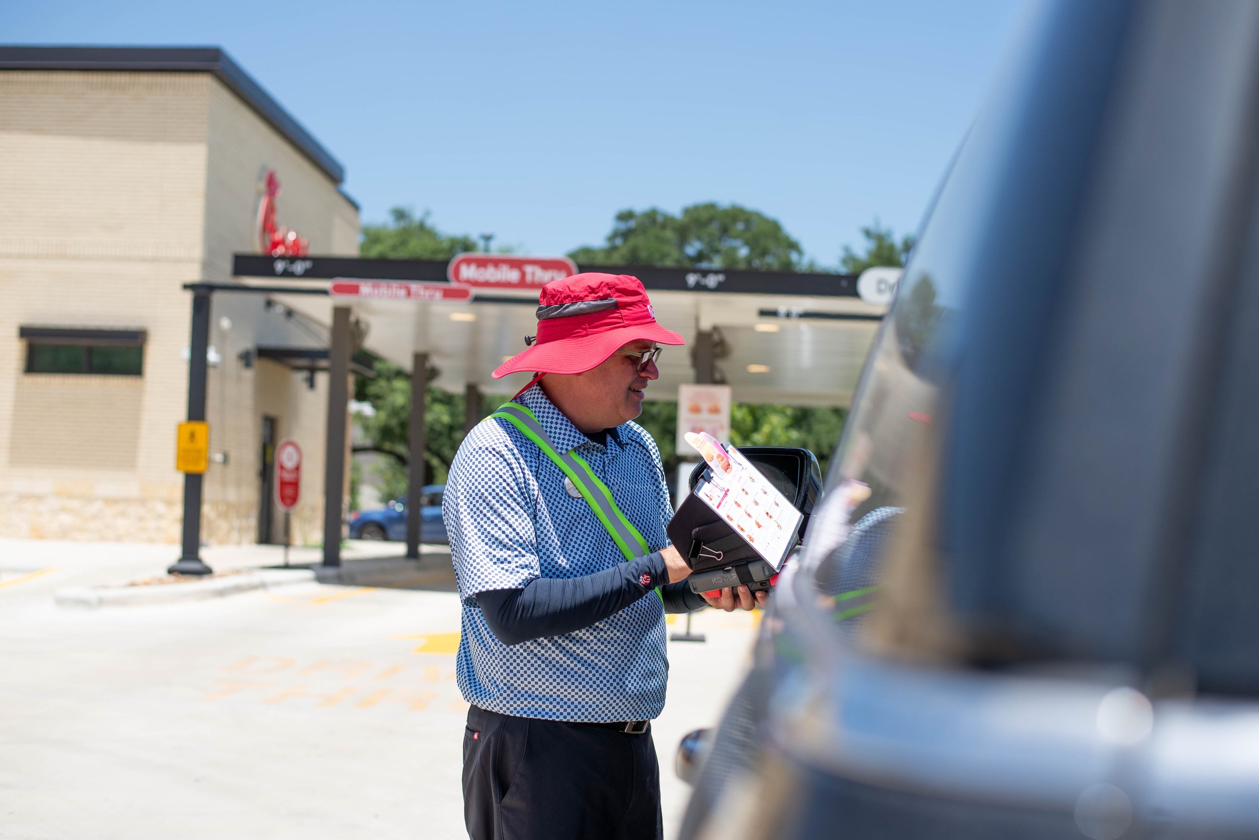 A man wearing a pink hat, glasses, and a blue checkered shirt with reflective vest is making a payment at a drive-thru window of a fast-food restaurant.