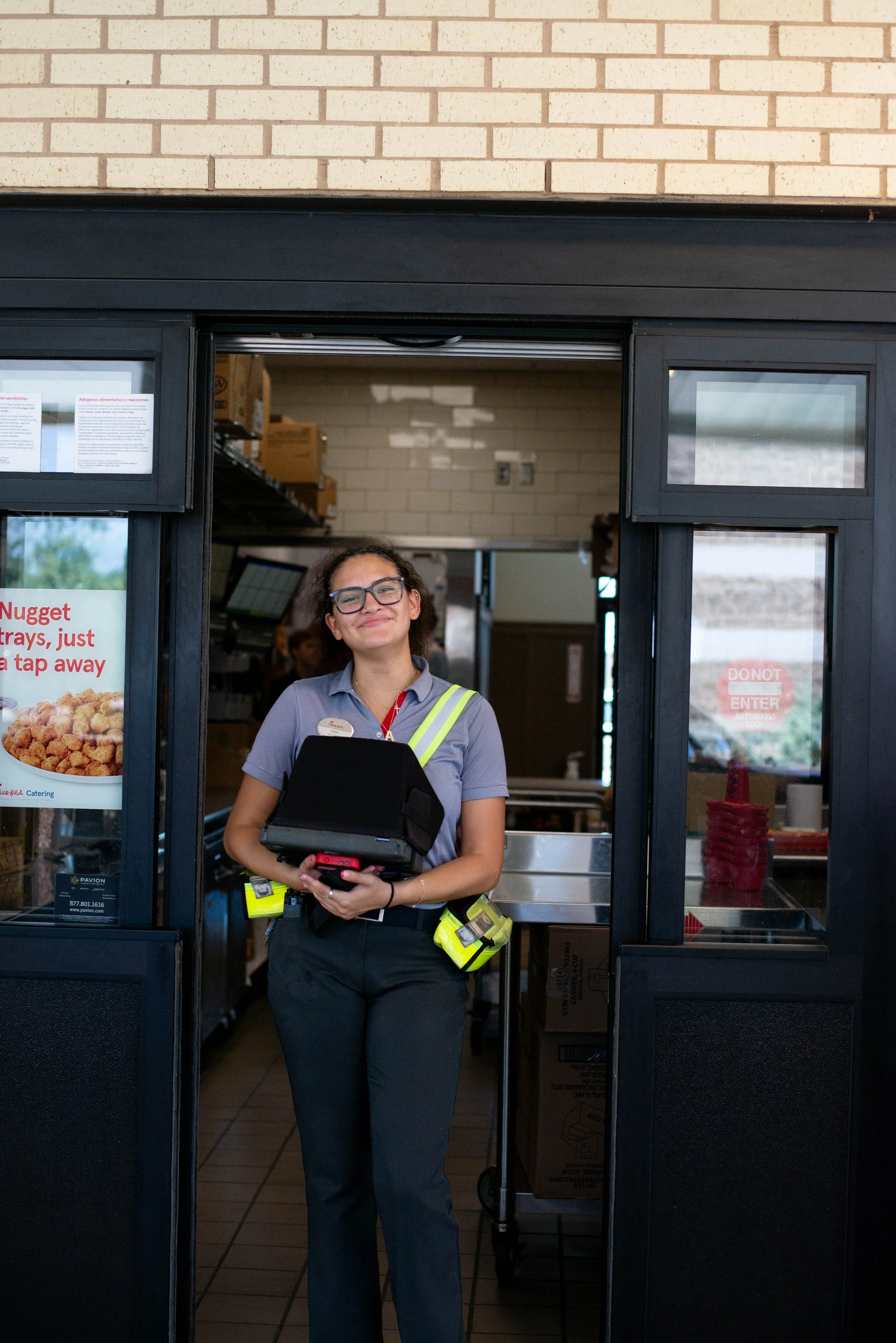 A smiling female employee in a Chick-fil-A uniform standing in a restaurant doorway, holding a digital order device with a yellow security badge clipped to her shirt, inside a fast-food restaurant.