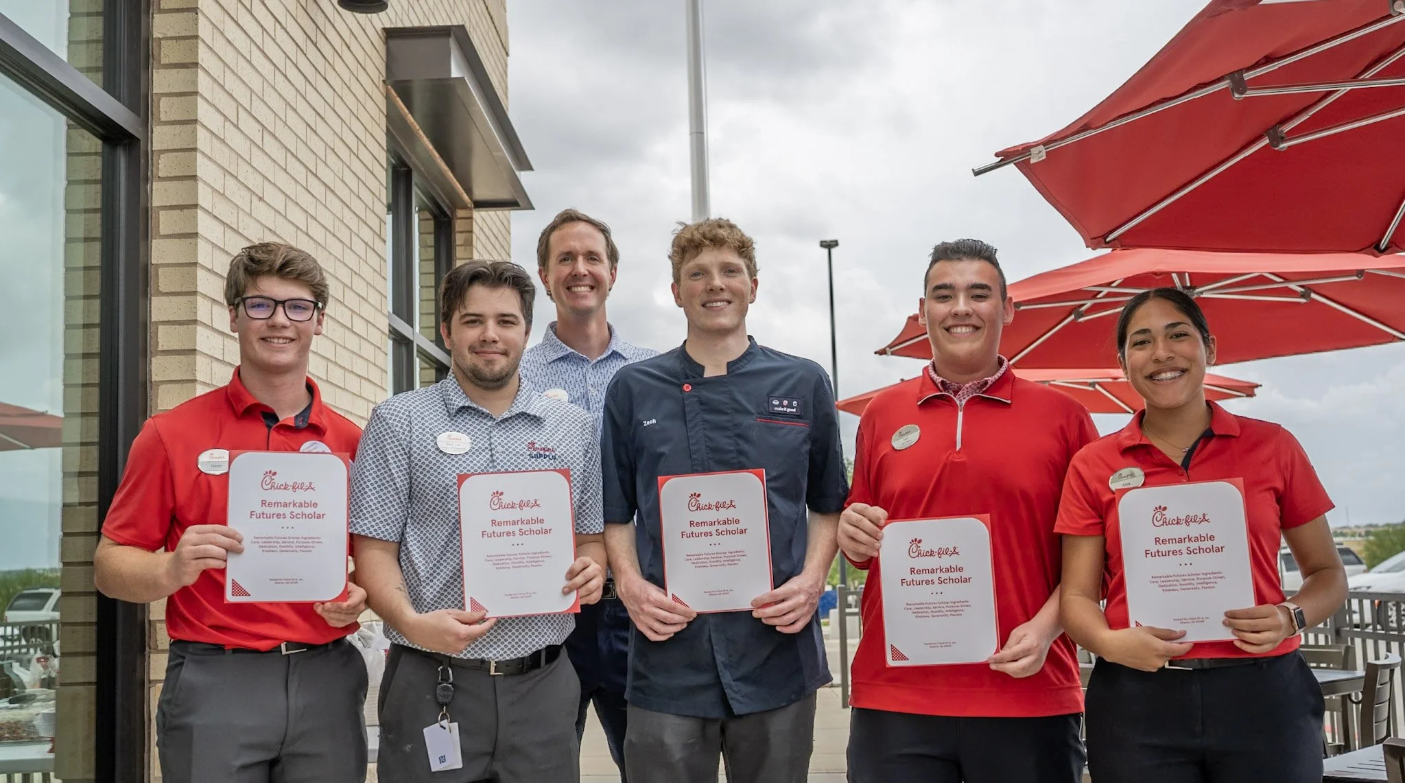 Group of six diverse young people standing outdoors, smiling, holding certificates that read 'Chick-fil-A Remarkable Futures Scholar,' in front of a building with red umbrellas overhead.