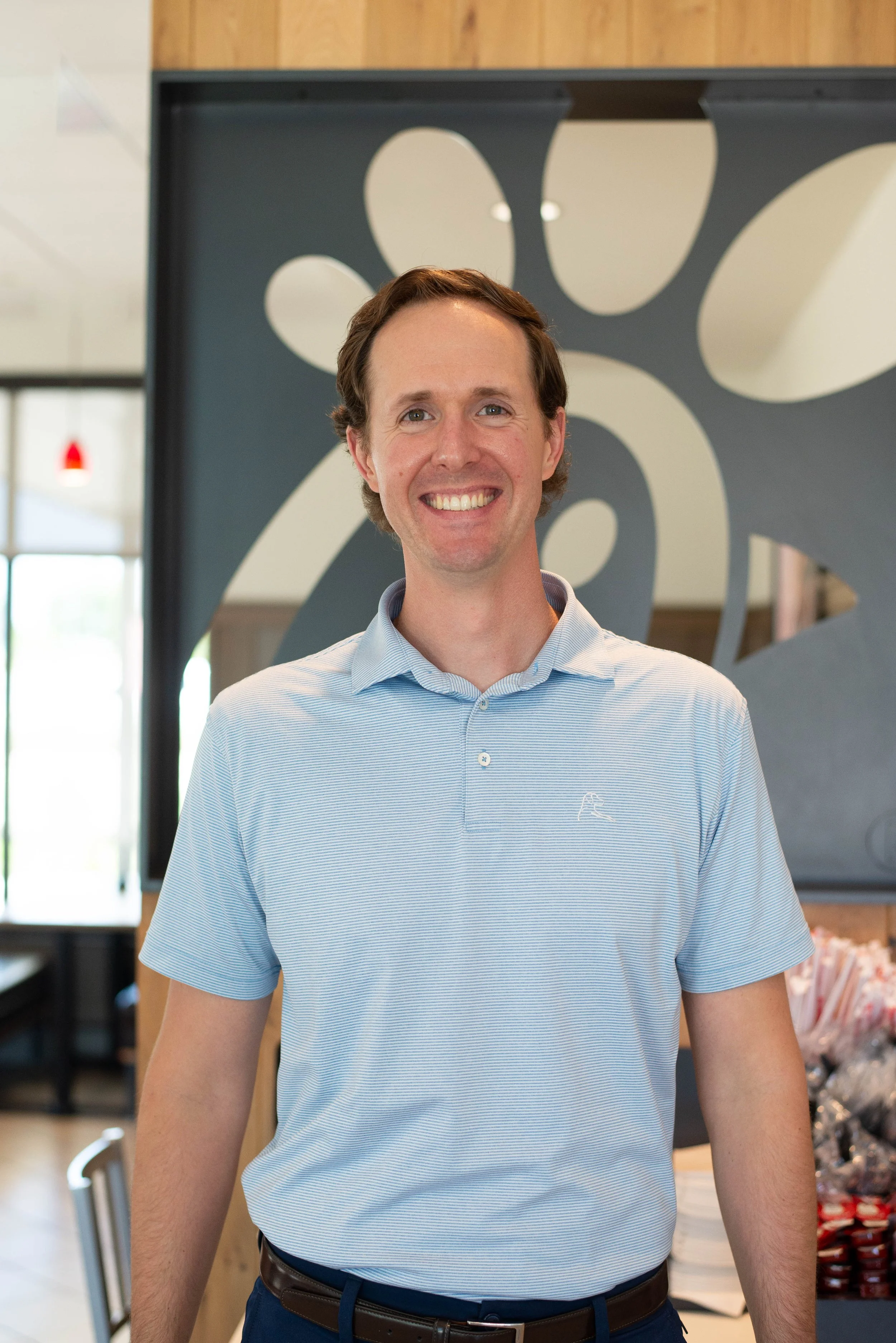 A smiling man wearing a light blue polo shirt standing inside a modern store or cafe. Background features a decorative black panel with abstract shapes and shelves with merchandise.
