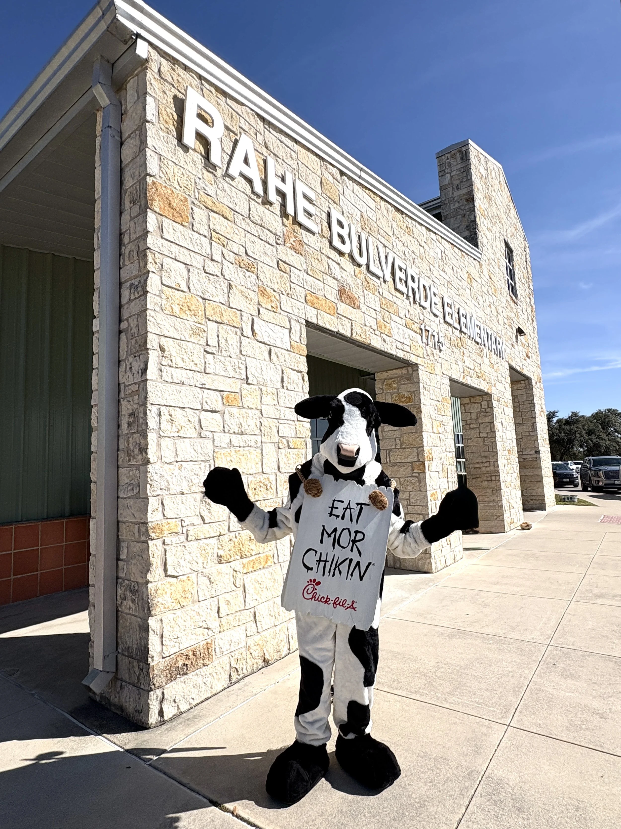 Person dressed in a cow costume holding a Chick-fil-A sign outside a building with stone walls and a sign that reads "RICHER BOULEVARD ELEMENTARY 171" on a sunny day.