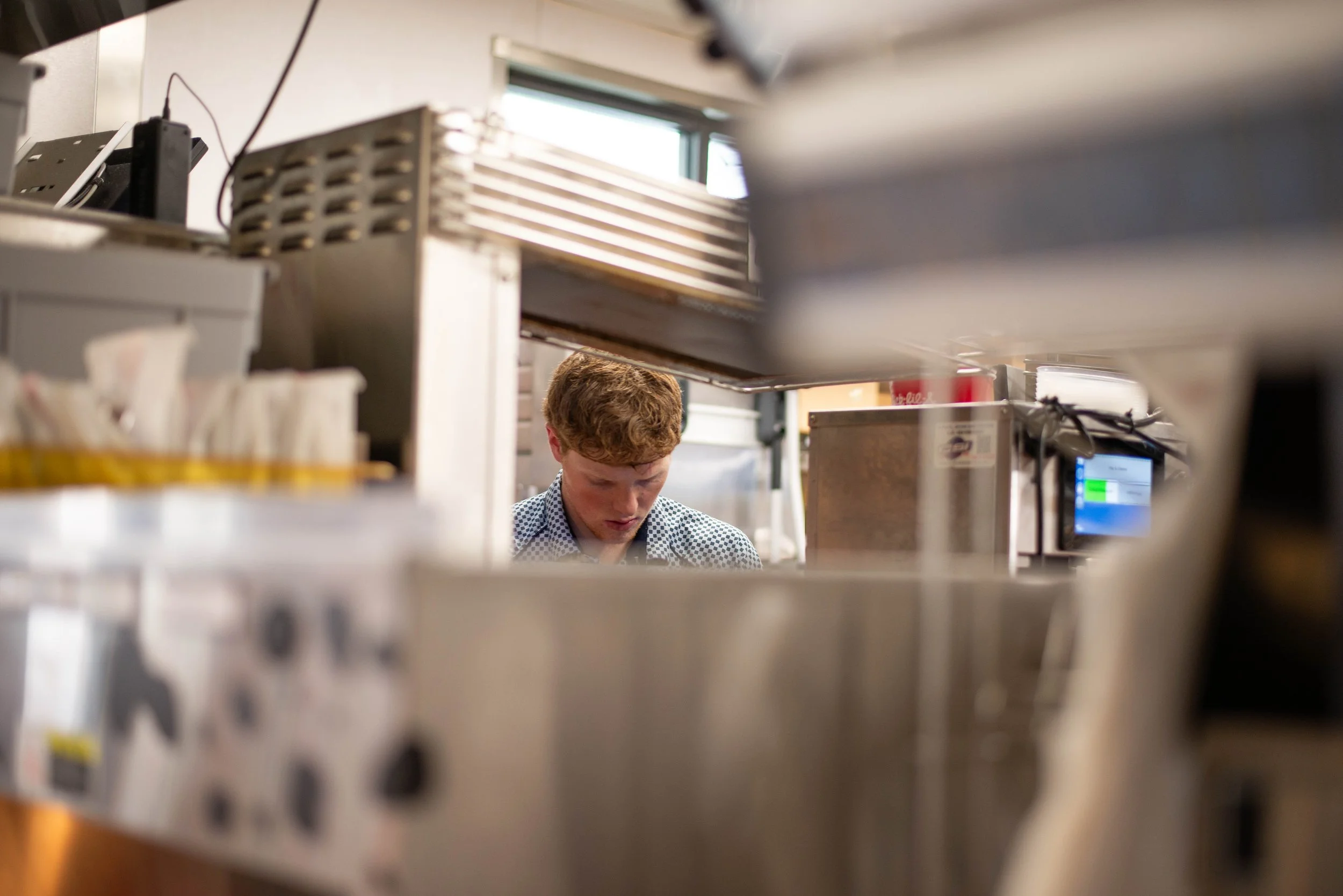 A person with curly hair wearing a patterned shirt is working or reading at a table in what appears to be a kitchen or food preparation area, with various equipment and supplies around, seen through a gap in a metal shelving unit.