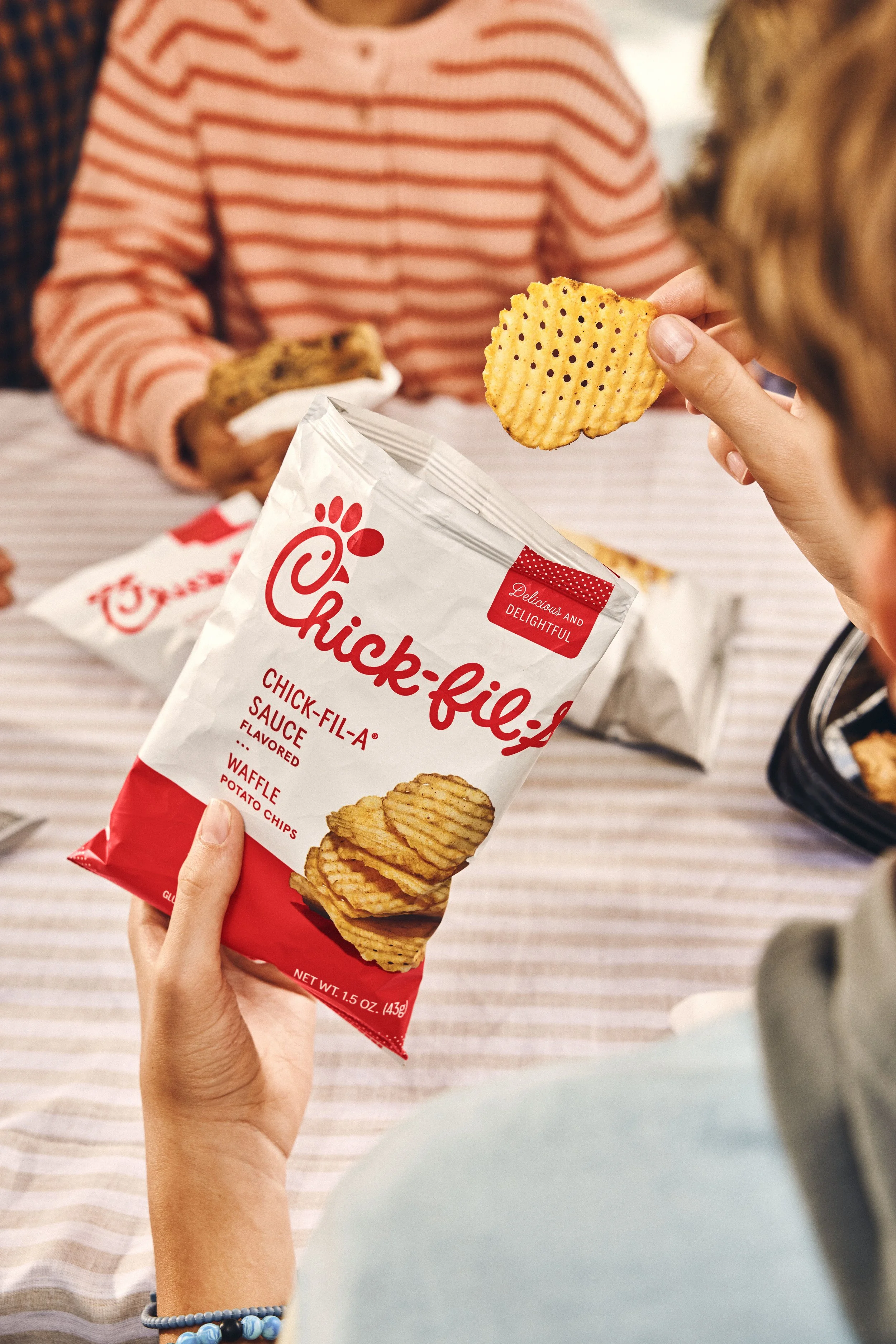 Person holding a bag of Chick-fil-A waffle potato chips with a person in the background wearing a pink and white striped shirt.