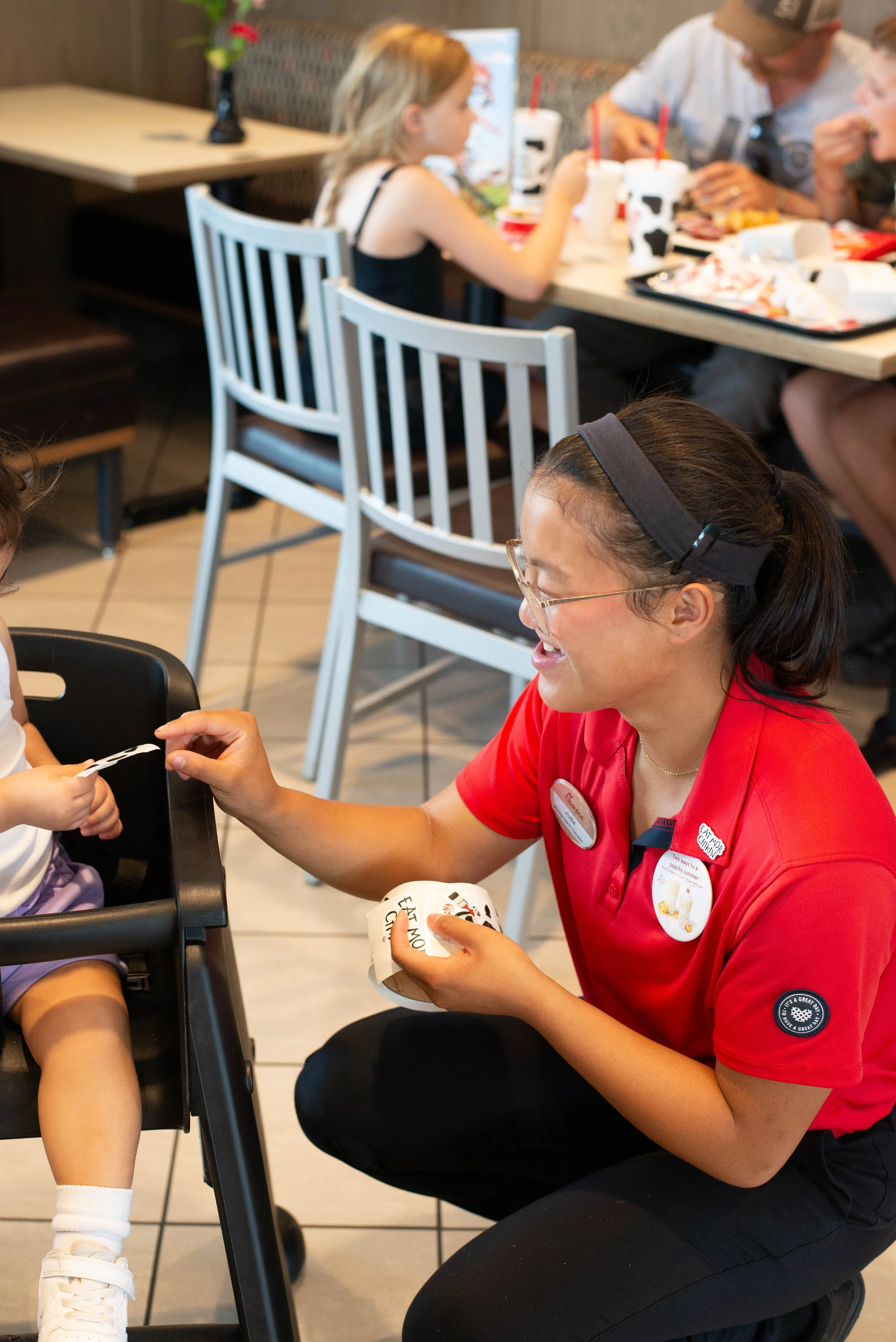 A woman in a red uniform talking to a young girl eating in a restaurant.