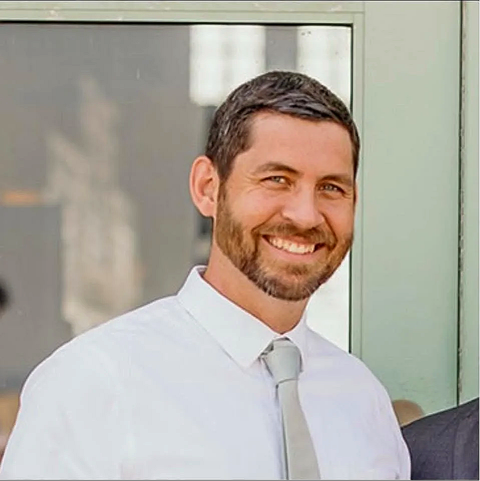 A smiling man with short dark hair and a beard, dressed in a white shirt and light gray tie, standing in front of a glass window or door.
