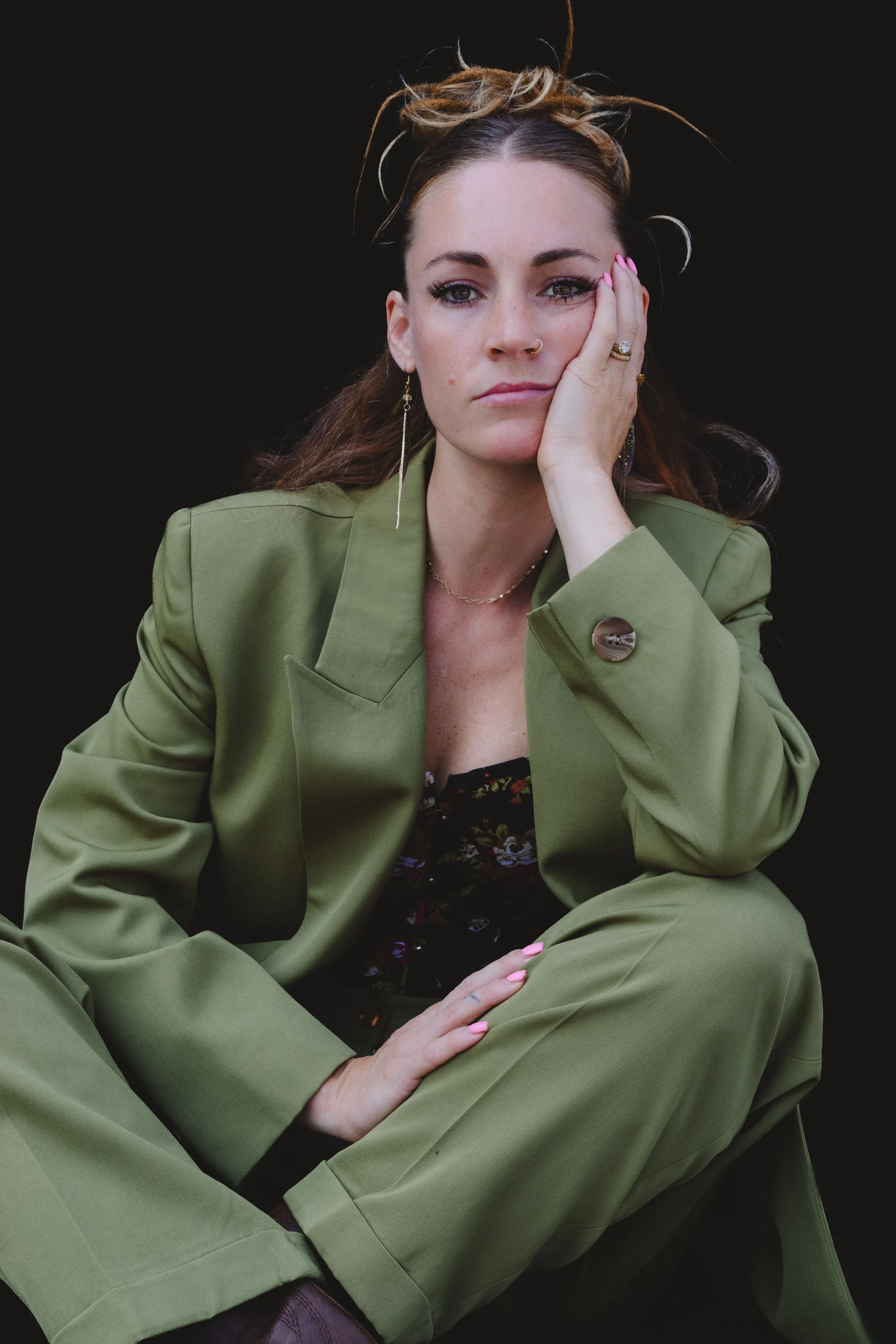 Woman in a green suit seated with her hand resting on her cheek in a moody studio portrait