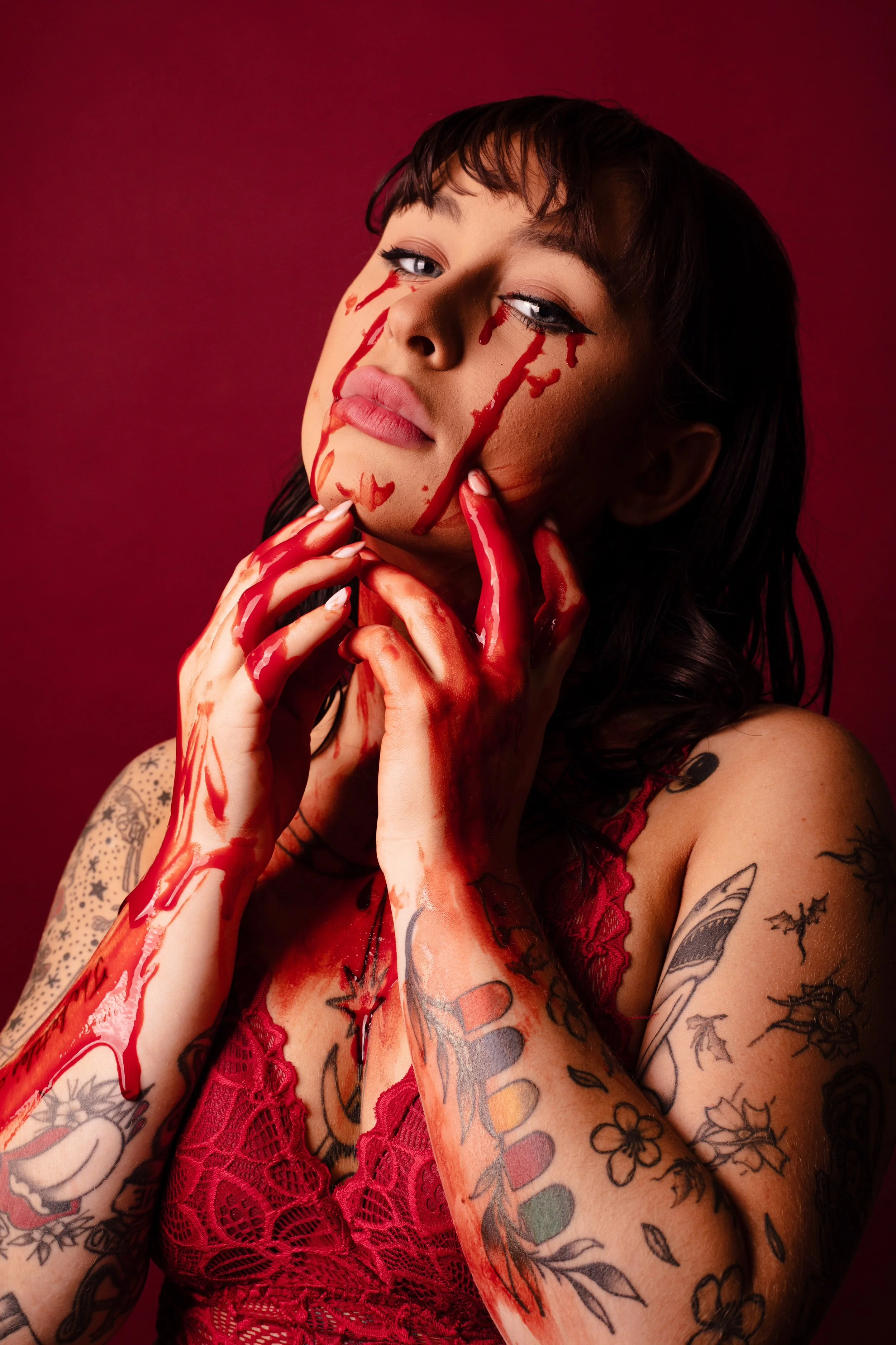 Tattooed woman with dramatic red paint on her face and hands against a red backdrop in a conceptual portrait