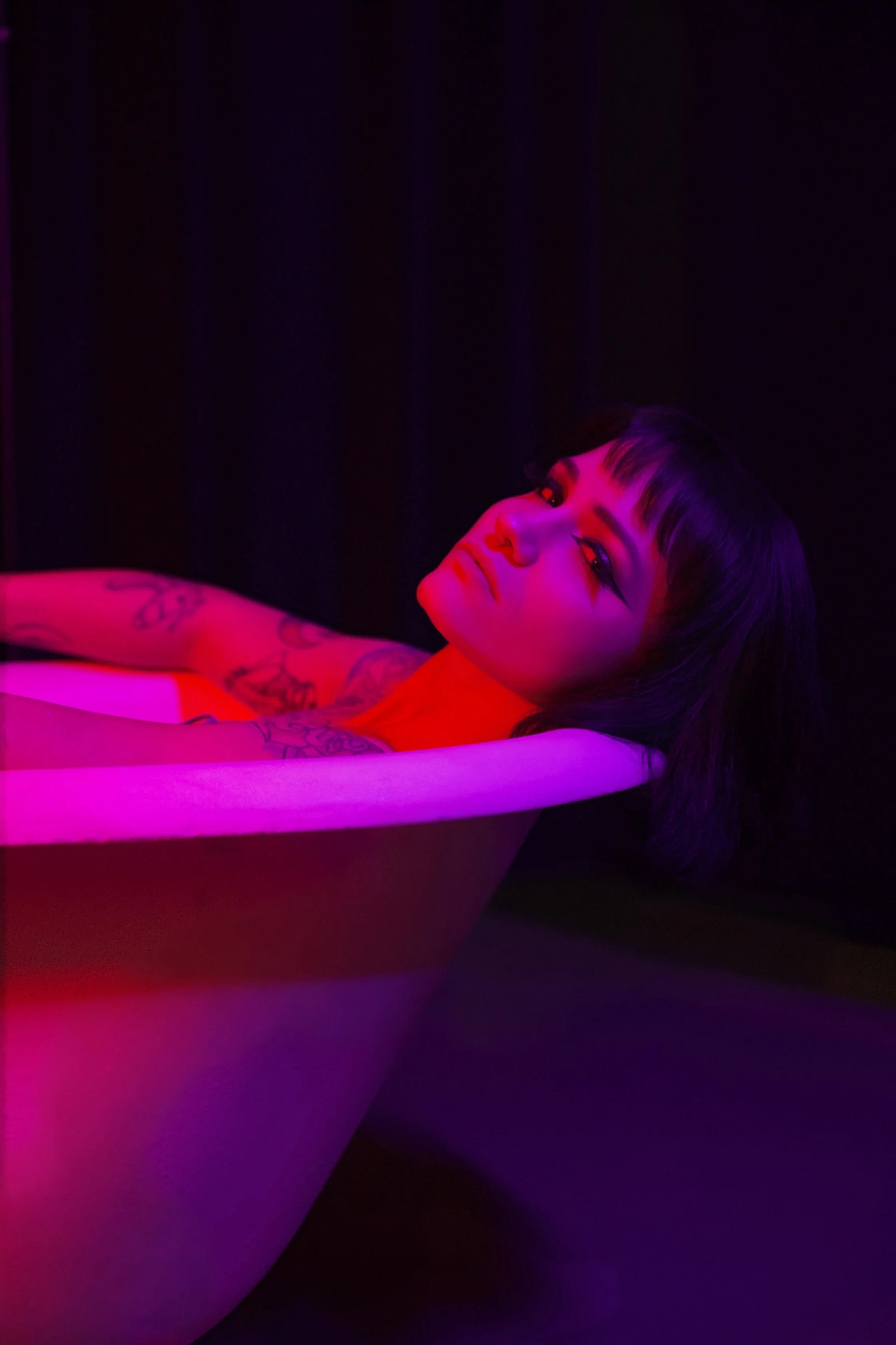 Tattooed woman relaxing in a bathtub lit with pink and purple neon light