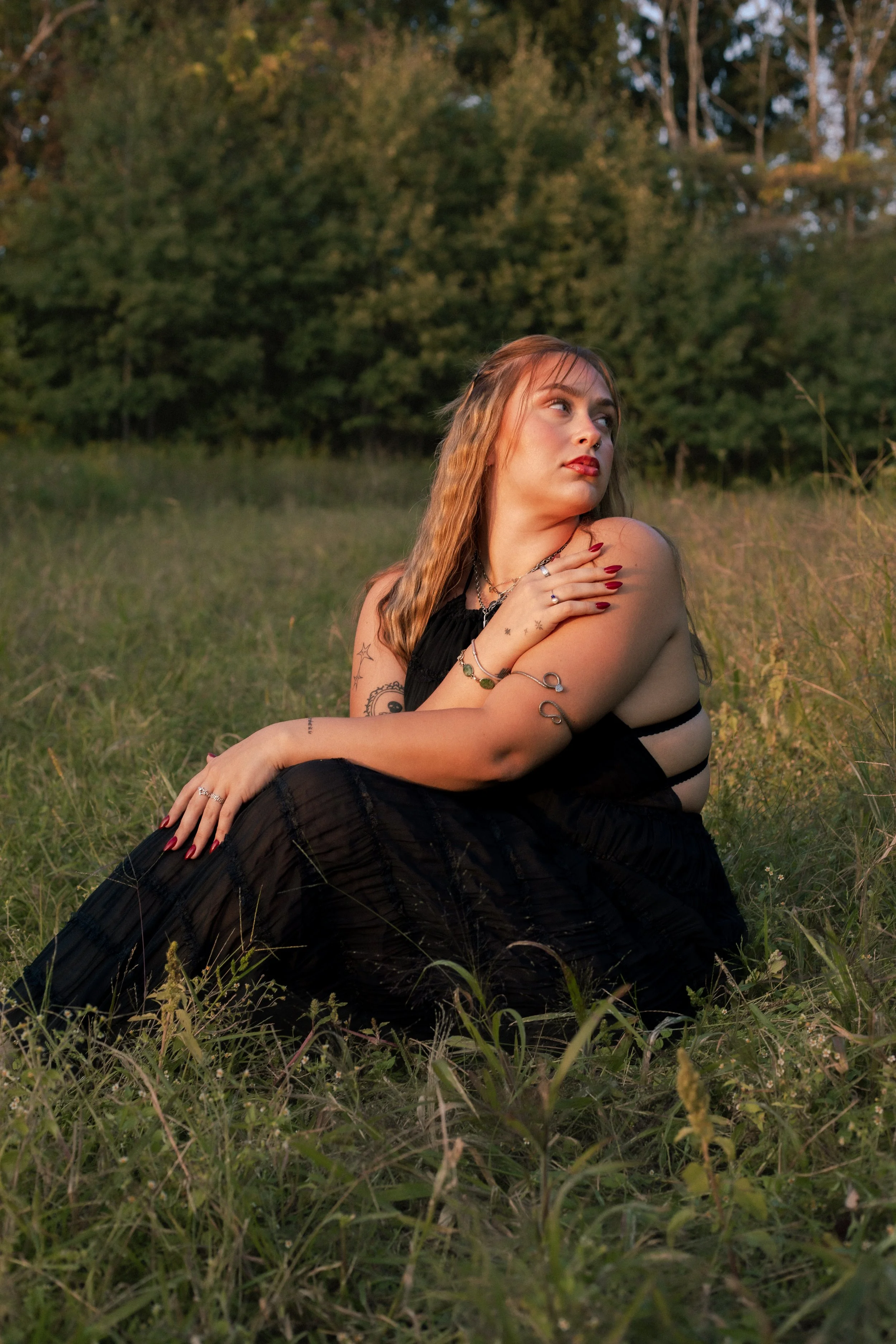 Woman in a black dress seated in tall grass at sunset during a golden hour portrait session in Utica NY