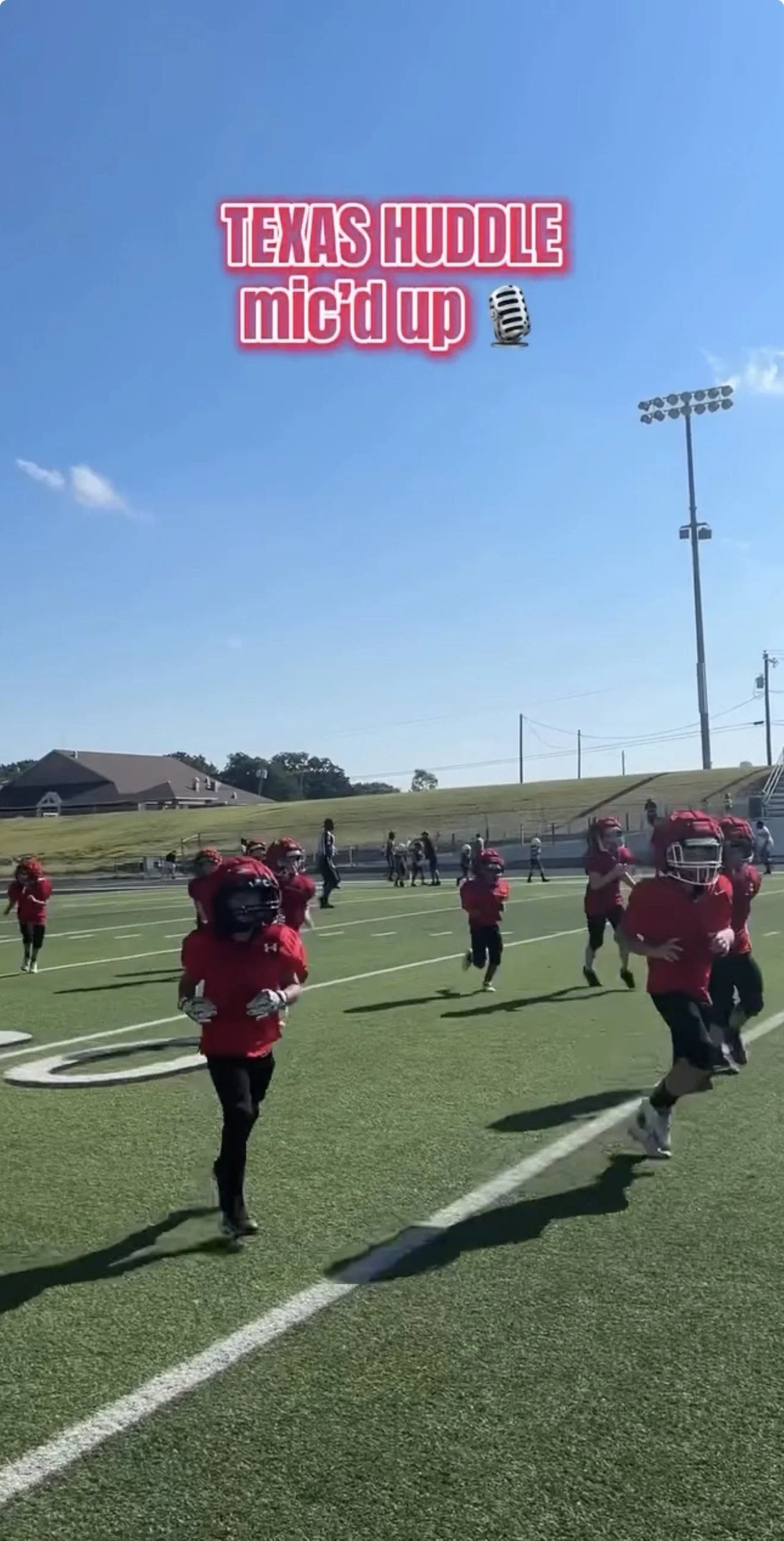 Young football players in red uniforms and helmets running on a field during a game or practice. A few adults are on the sidelines, and there are stadium lights and houses in the background on a clear, sunny day.