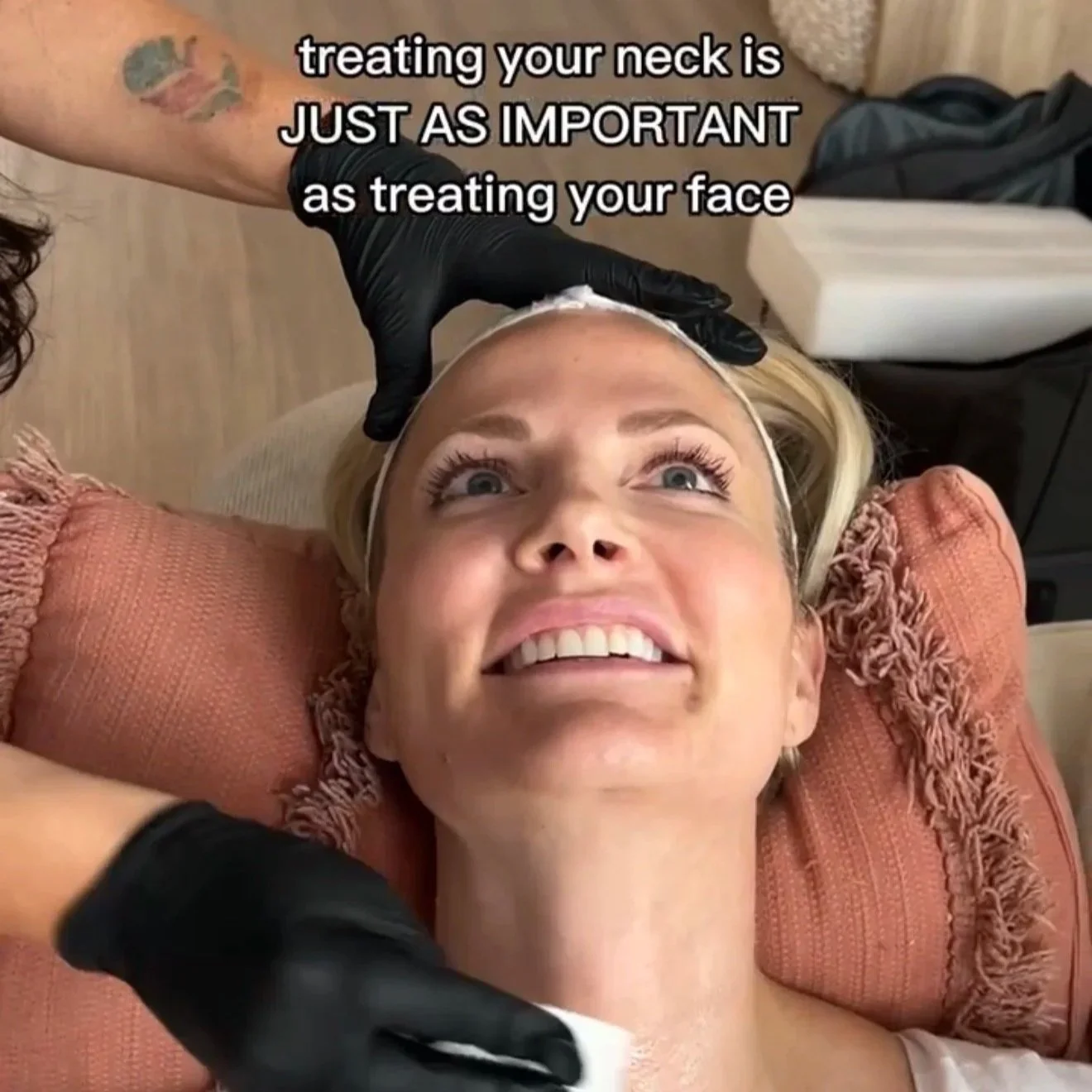 A woman receiving a neck treatment at a spa or salon, lying back on a pillow with a smiling expression, while a professional wearing black gloves applies treatment to her neck.