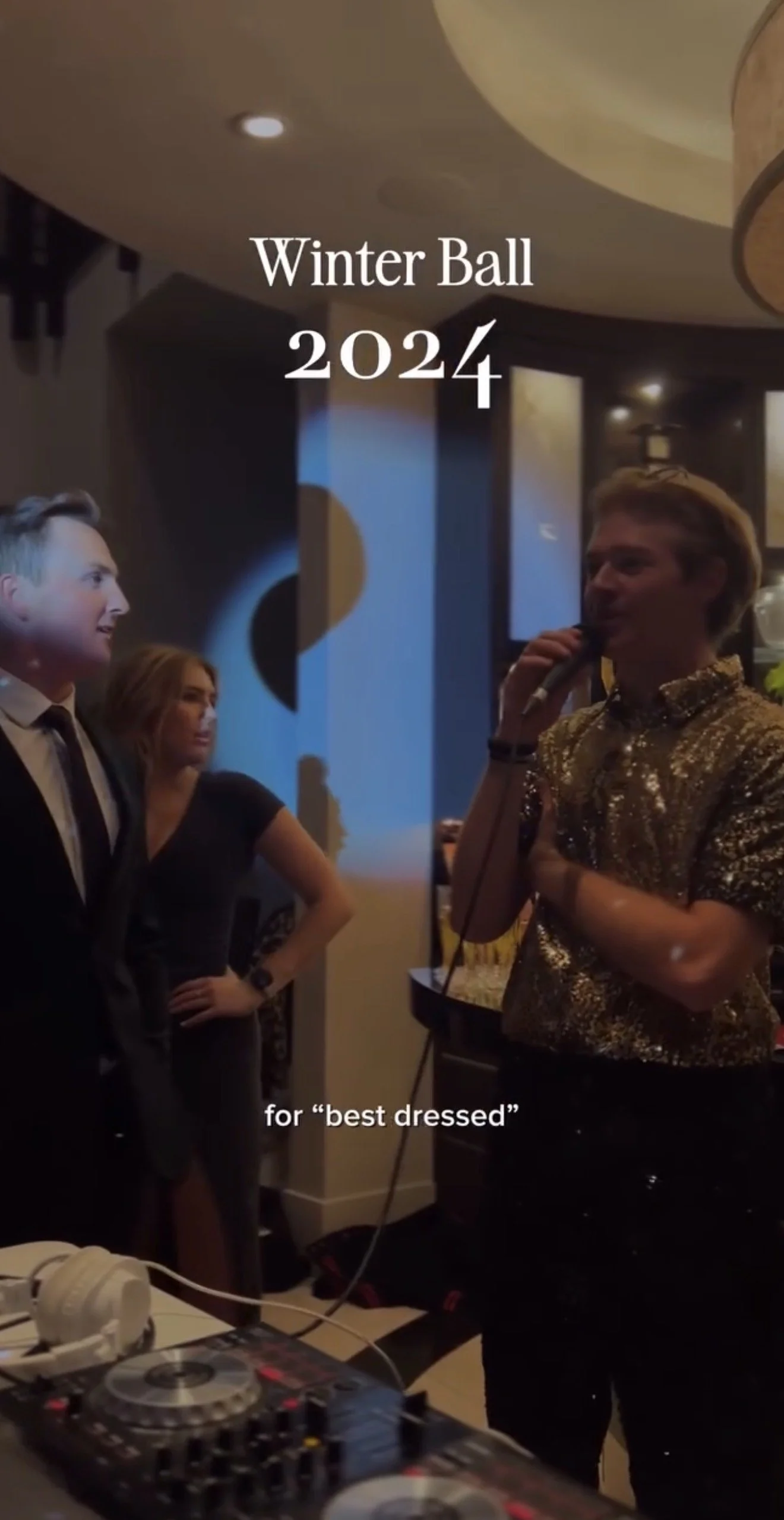 A group of people at a winter event with a DJ booth in the foreground. One person is speaking into a microphone, wearing a shiny, gold, sequined shirt, while the others are listening. Text on the image reads "Winter Ball 2024" and "for 'best dressed'