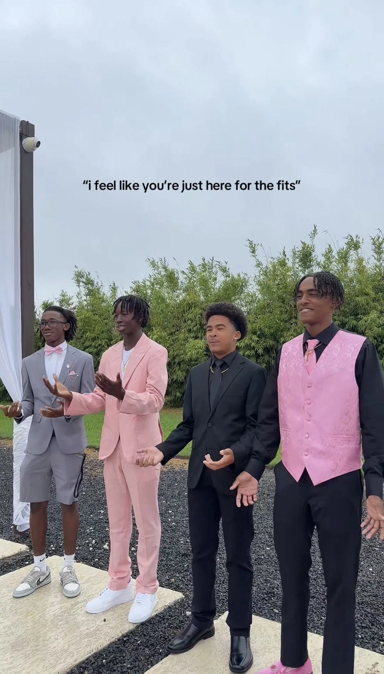 Four young men dressed in formal suits, standing outdoors in front of green bushes, with a cloudy sky above. They are participating in a prayer or blessing, with hands open and eyes closed or looking down. One of them is wearing a pink vest and pink 