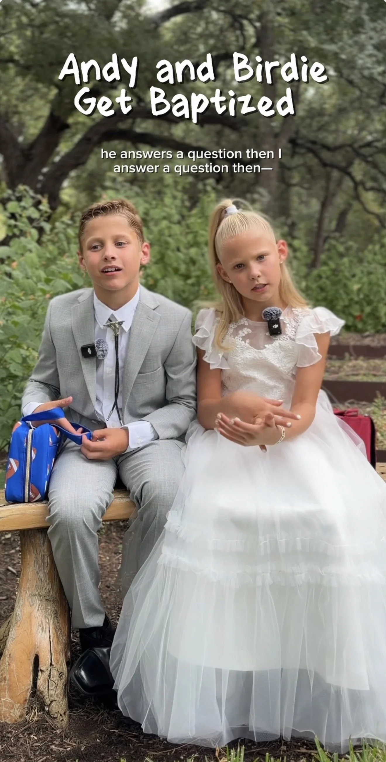 Two young children, a boy and a girl, sitting on a wooden bench in a park. The boy is dressed in a light gray suit with a white shirt and a bolo tie, holding a blue school bag. The girl is wearing a white dress with lace and tulle, and has a micropho