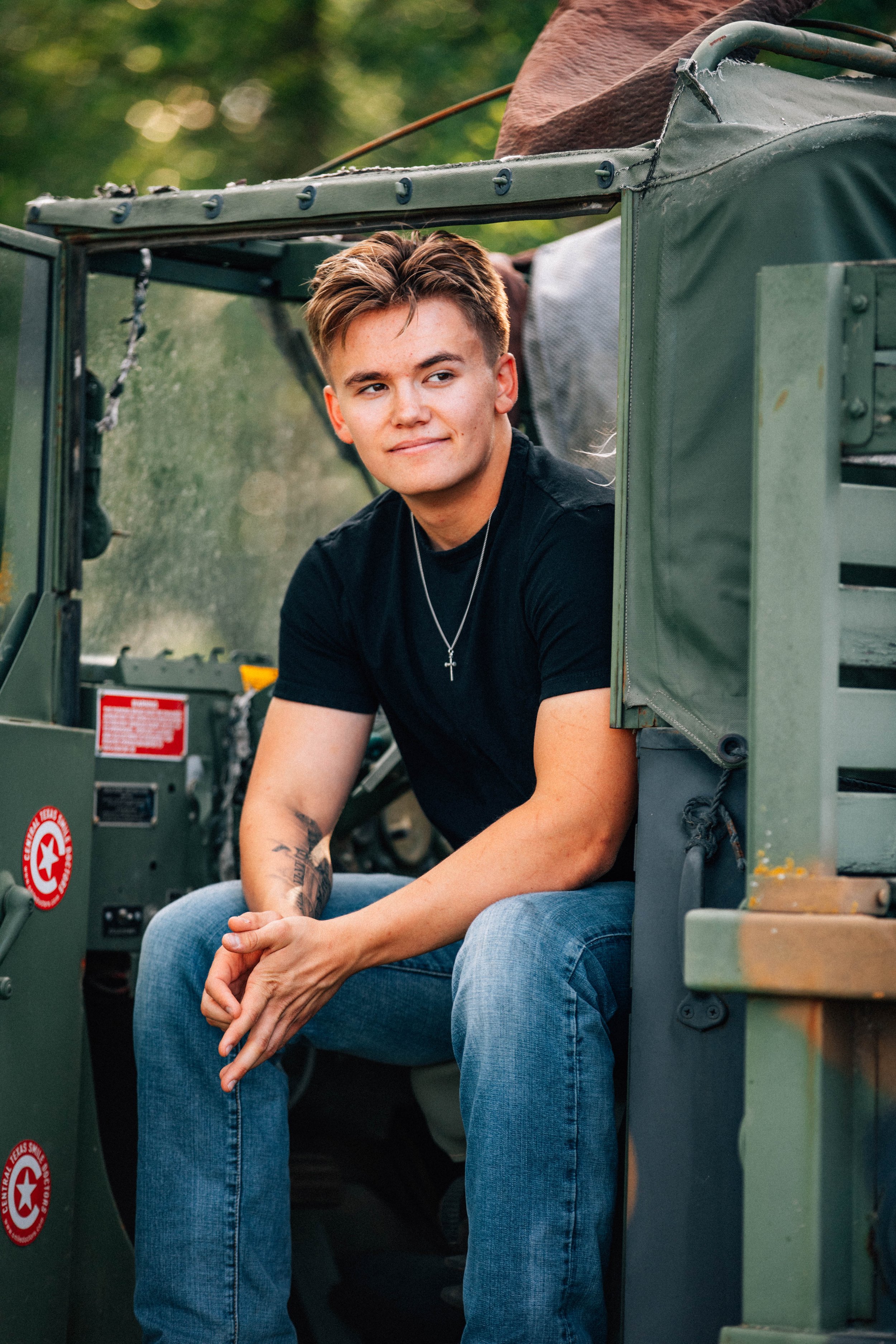 Young man sitting in a military vehicle, wearing a black t-shirt, light blue jeans, with tattoos on his arm, and a silver cross necklace, outdoors with green blurred background.