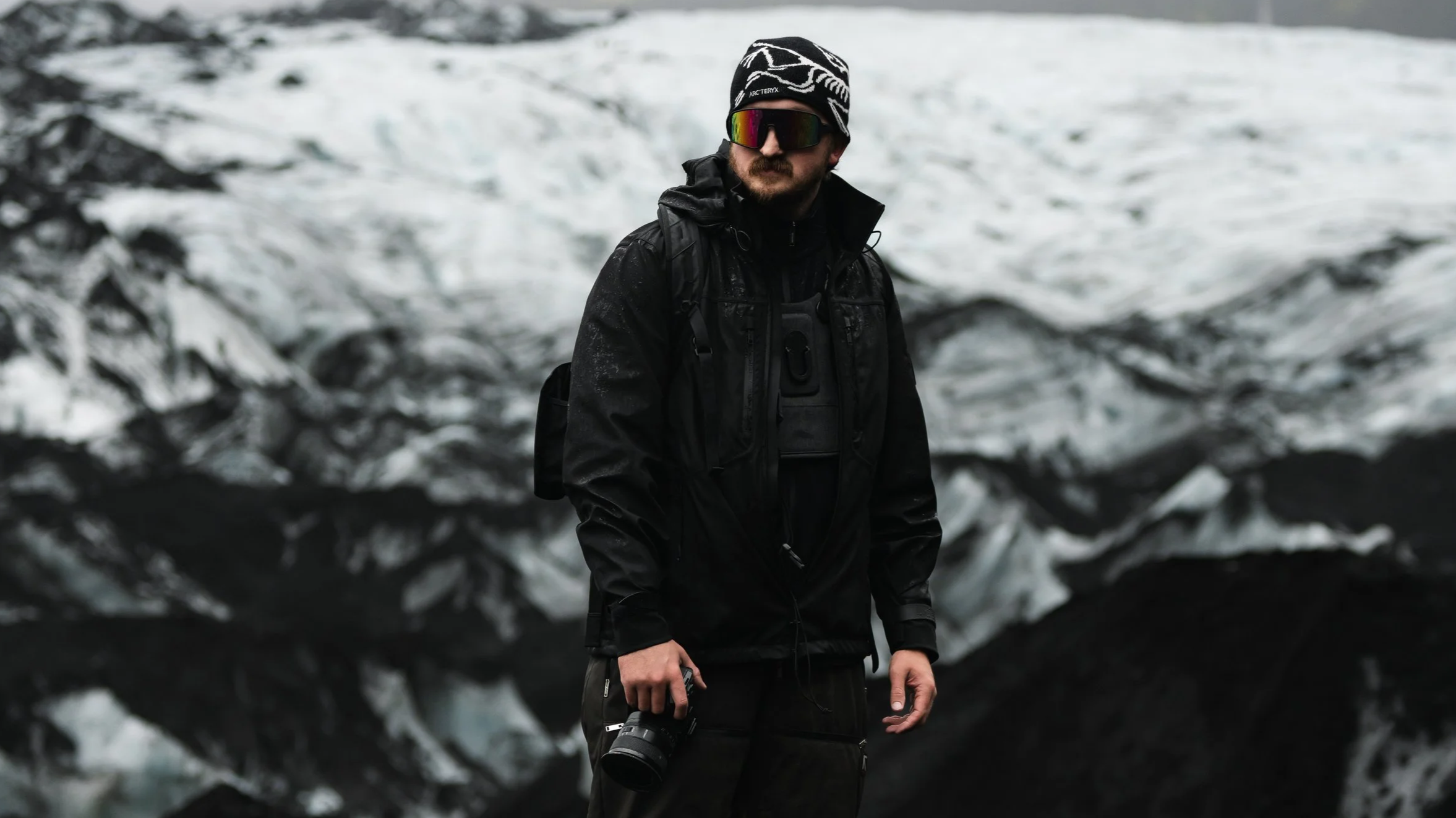 Man dressed in black outdoor gear, wearing sunglasses and a beanie, standing near a glacier in a cold environment.