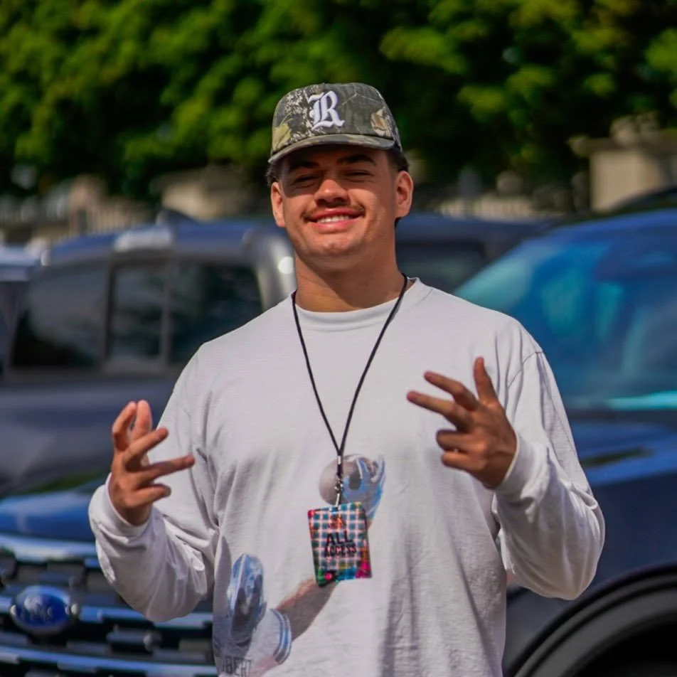 A young man smiling and making peace signs with his hands outdoors, wearing a camouflage cap, a white long-sleeve shirt, and a lanyard with a colorful patterned badge.