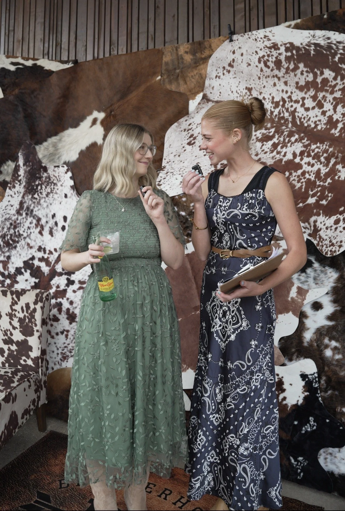 Two women in dresses talking and smiling indoors, with a backdrop of cowhide-patterned wallpaper and a vintage armchair.