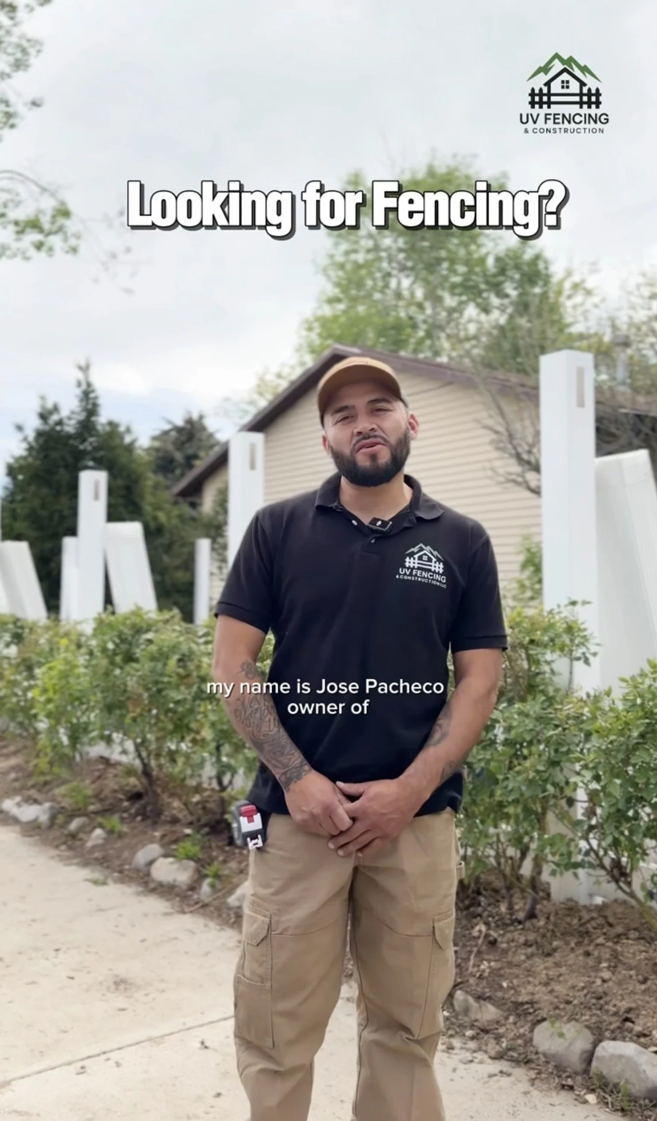 A man with a beard and tattoos wearing a black UV fencing T-shirt and tan cargo pants stands outdoors in front of a garden with weeds, shrubs, and white fencing posts. The sky is cloudy and trees are in the background.