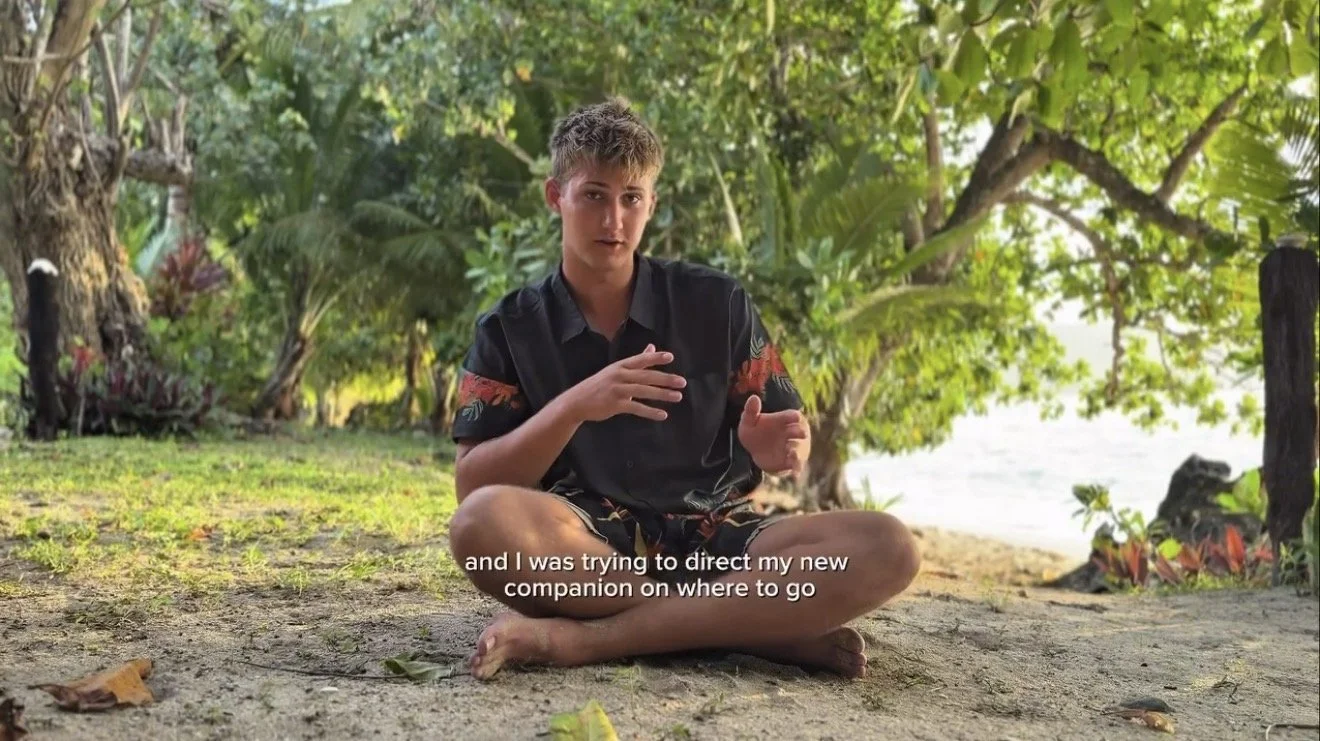 A young man with short light brown hair sitting cross-legged on the ground outdoors, surrounded by lush green trees and plants, with a beach or water body in the background, wearing a black shirt with red and yellow sleeves, speaking and gesturing wi