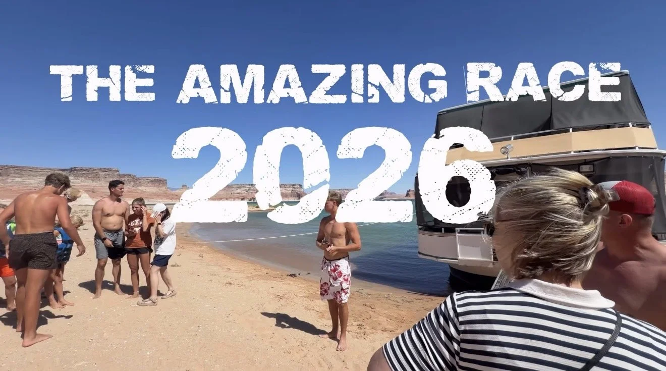 People gather on a sandy beach near a boat under a clear blue sky, with cliffs in the background. Overlaid text reads "The Amazing Race 2026."