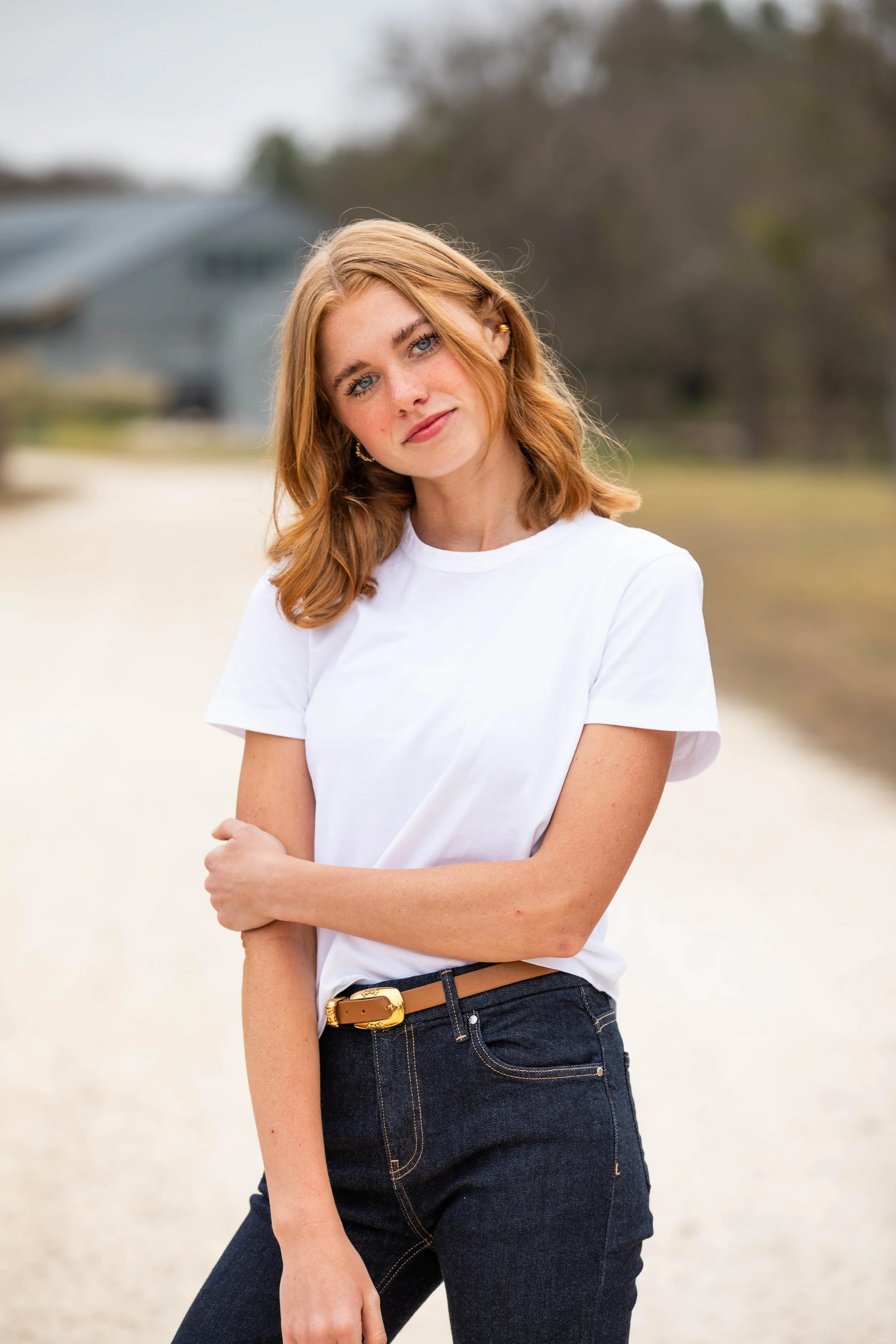 Young woman with red hair and blue eyes standing outdoors, wearing a white t-shirt and dark jeans, with a blurred background of trees and a house.