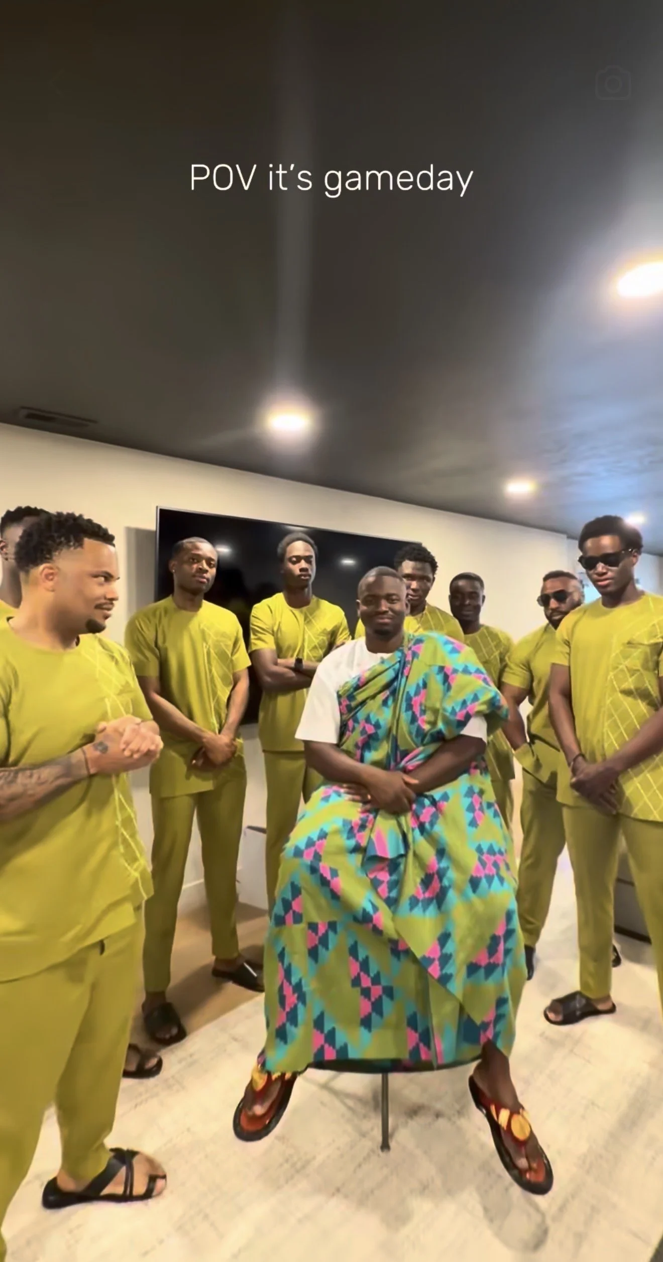 Group of people wearing yellow outfits, one person in colorful traditional attire, standing in a room with white walls and a black TV in the background.