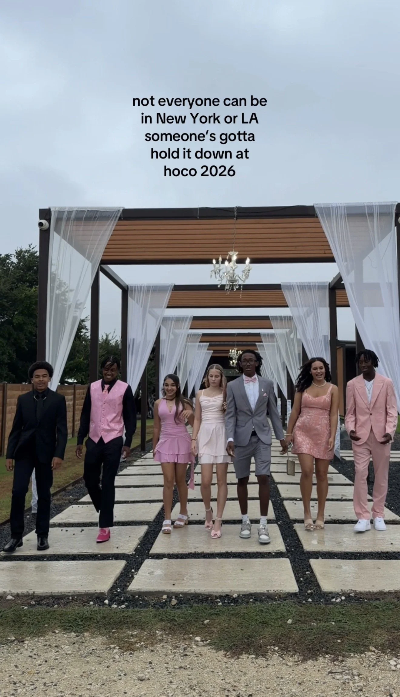 A group of young people dressed in formal attire walking outdoors on a path, with a decorative pergola structure overhead and text overlay indicating a message about future plans and a music festival in 2026.