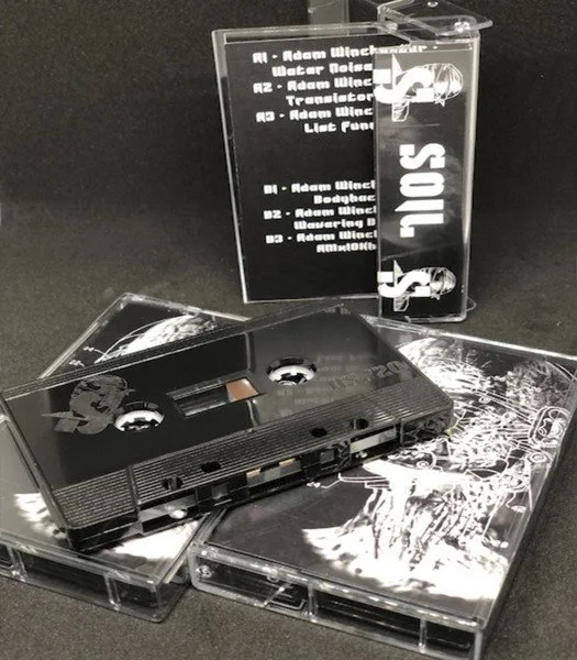 A black audio cassette tape, a clear plastic case with a list of song titles, and two cassette tape cases with artwork, all arranged on a black surface.
