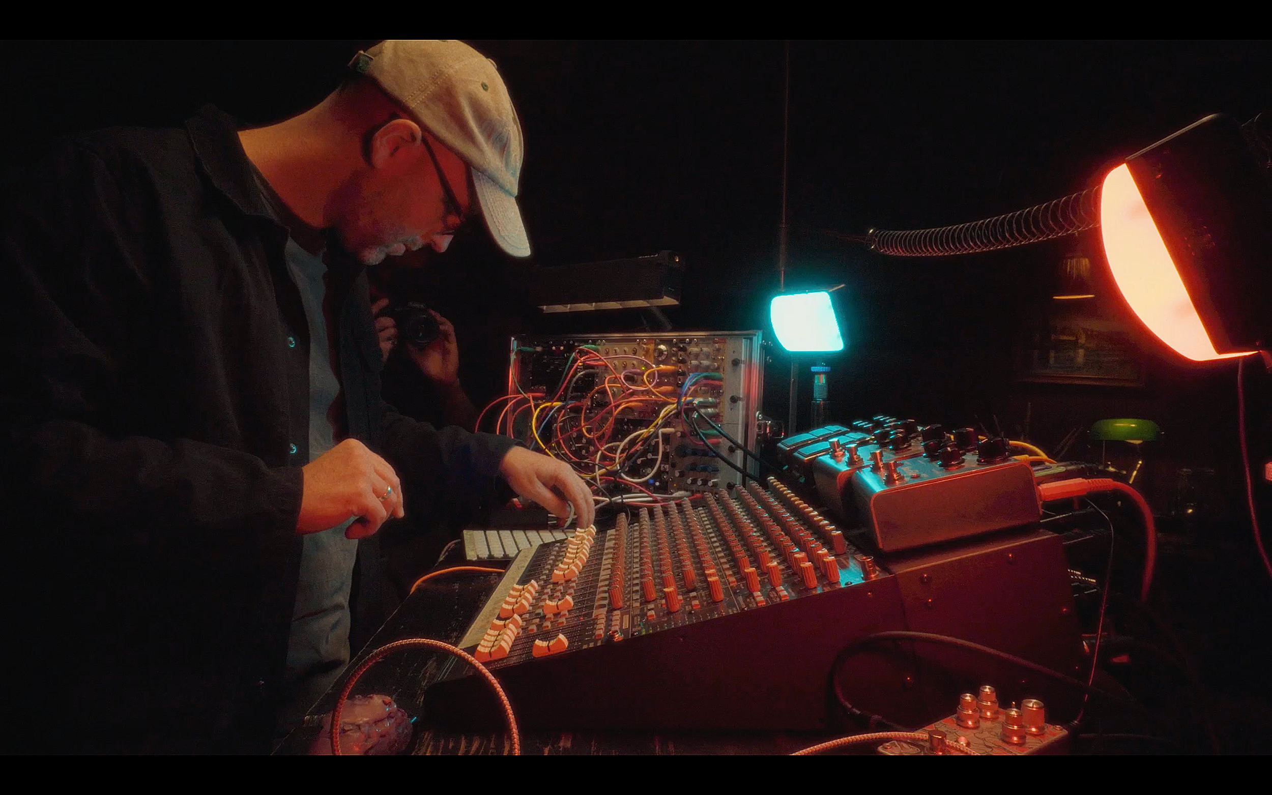 A person wearing a beige cap and glasses operates electronic music equipment, including a mixer with numerous wires, knobs, and buttons, in a dark room illuminated by colorful LED lights.