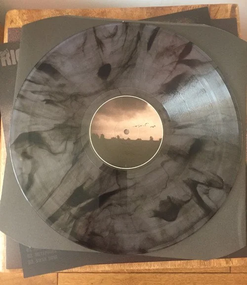 A black vinyl record with black and white smoke-like patterns on it, placed on top of a black cover sleeve.