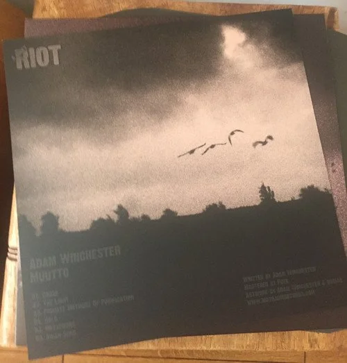 Black and white photograph of a cloudy sky with three birds flying, landscape with trees and hills, and text indicating it's an album cover for Riot, with Adam Winchester Muutto's name.