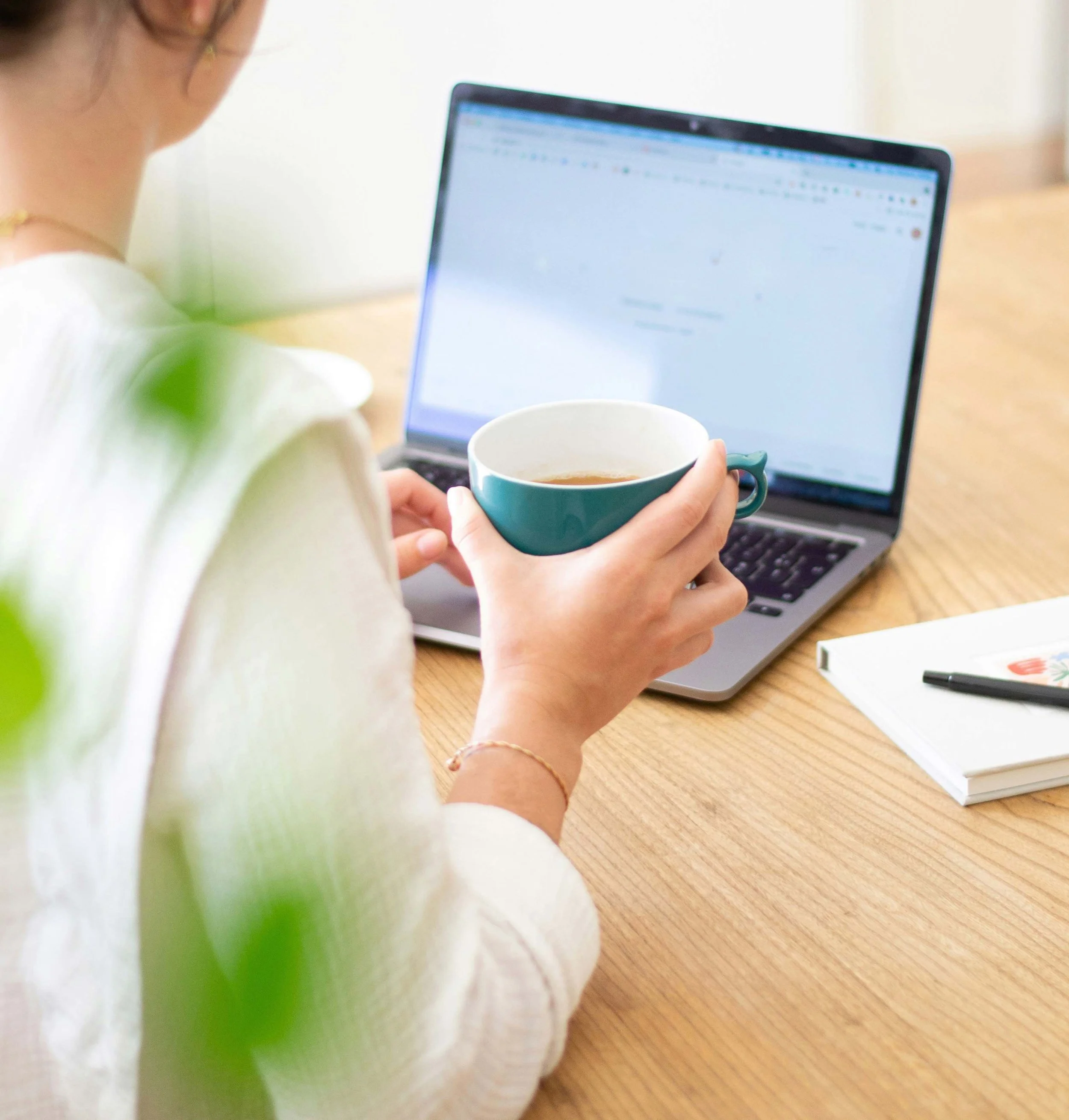 A person holding a cup of tea while working on a laptop at a wooden desk.