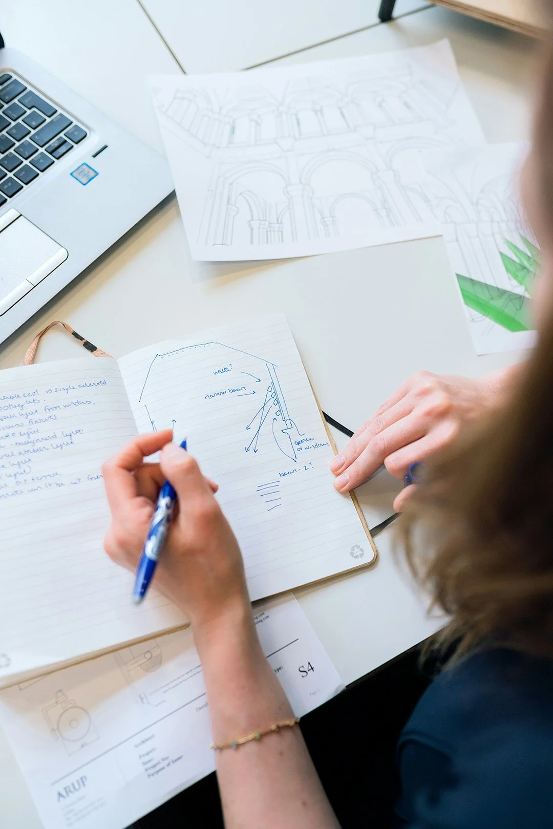 Person taking notes in a notebook with sketches and handwritten notes, surrounded by architectural drawings and a laptop on a white desk.