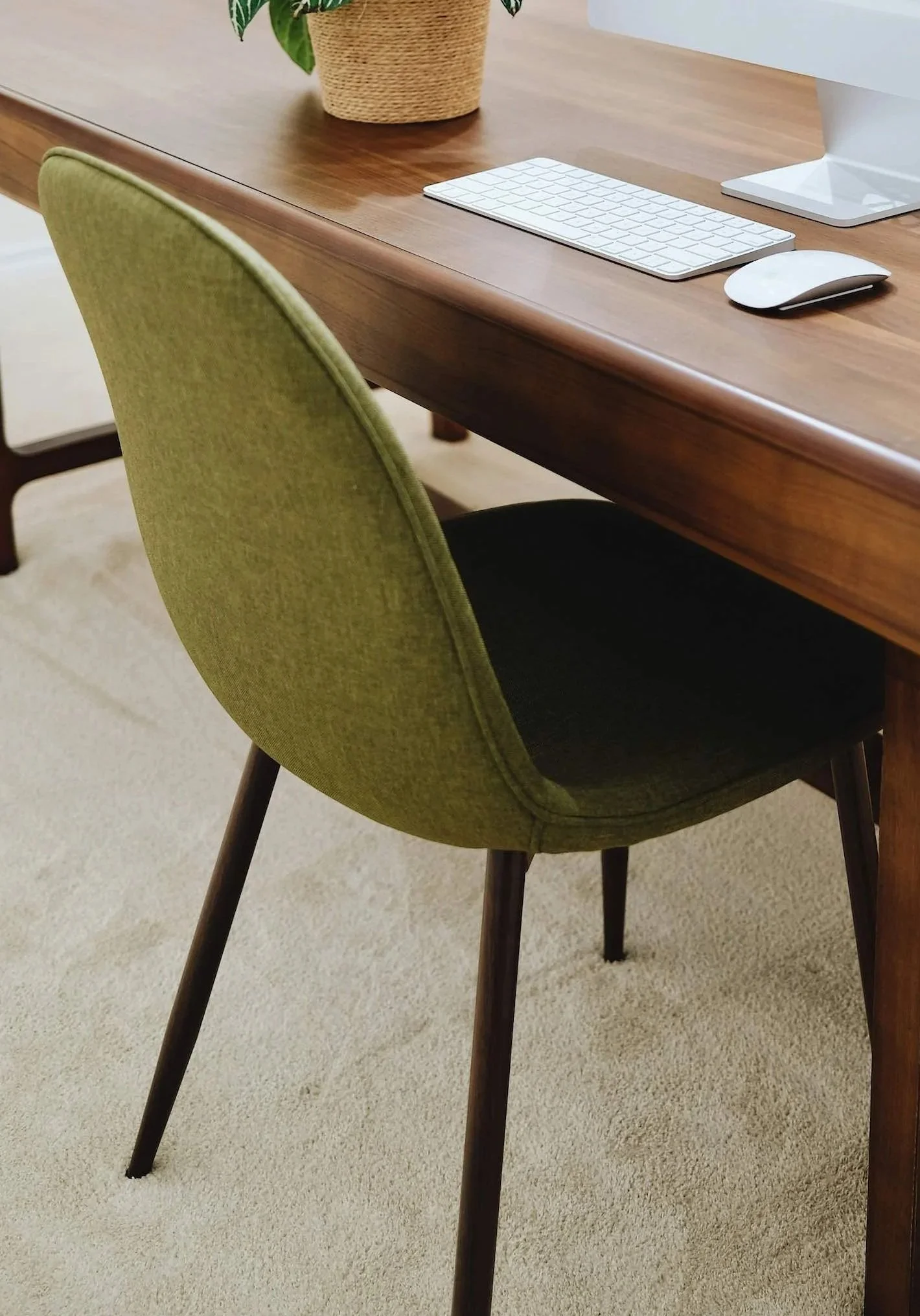 A wooden table with a green upholstered chair, a computer monitor, a white keyboard, a white mouse, and a potted plant in a woven basket on top.