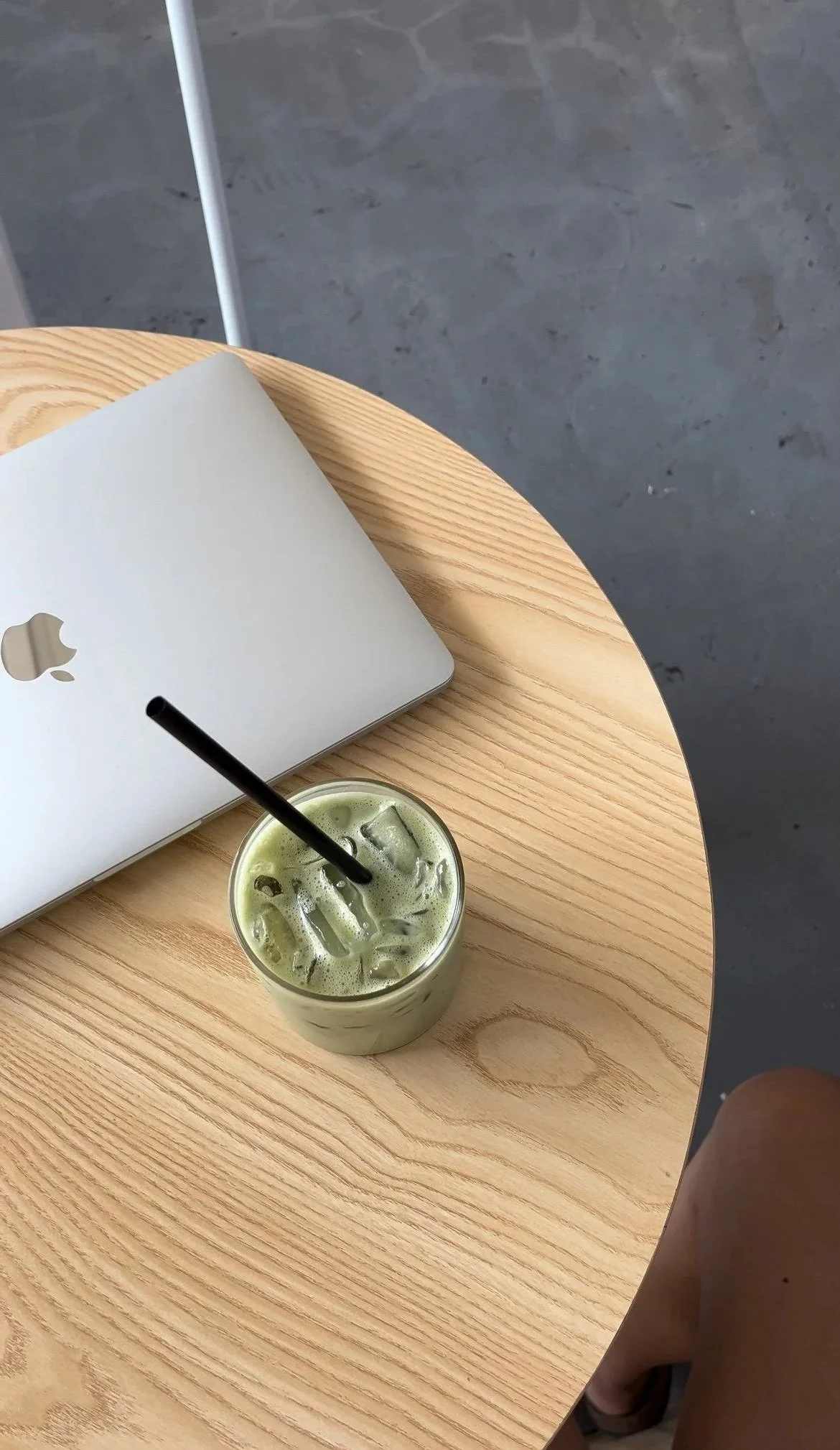Top view of a round wooden table with a closed MacBook, a glass of iced green drink with ice cubes and a black straw, and part of a person's leg and foot wearing a sandal. The background shows a gray floor.