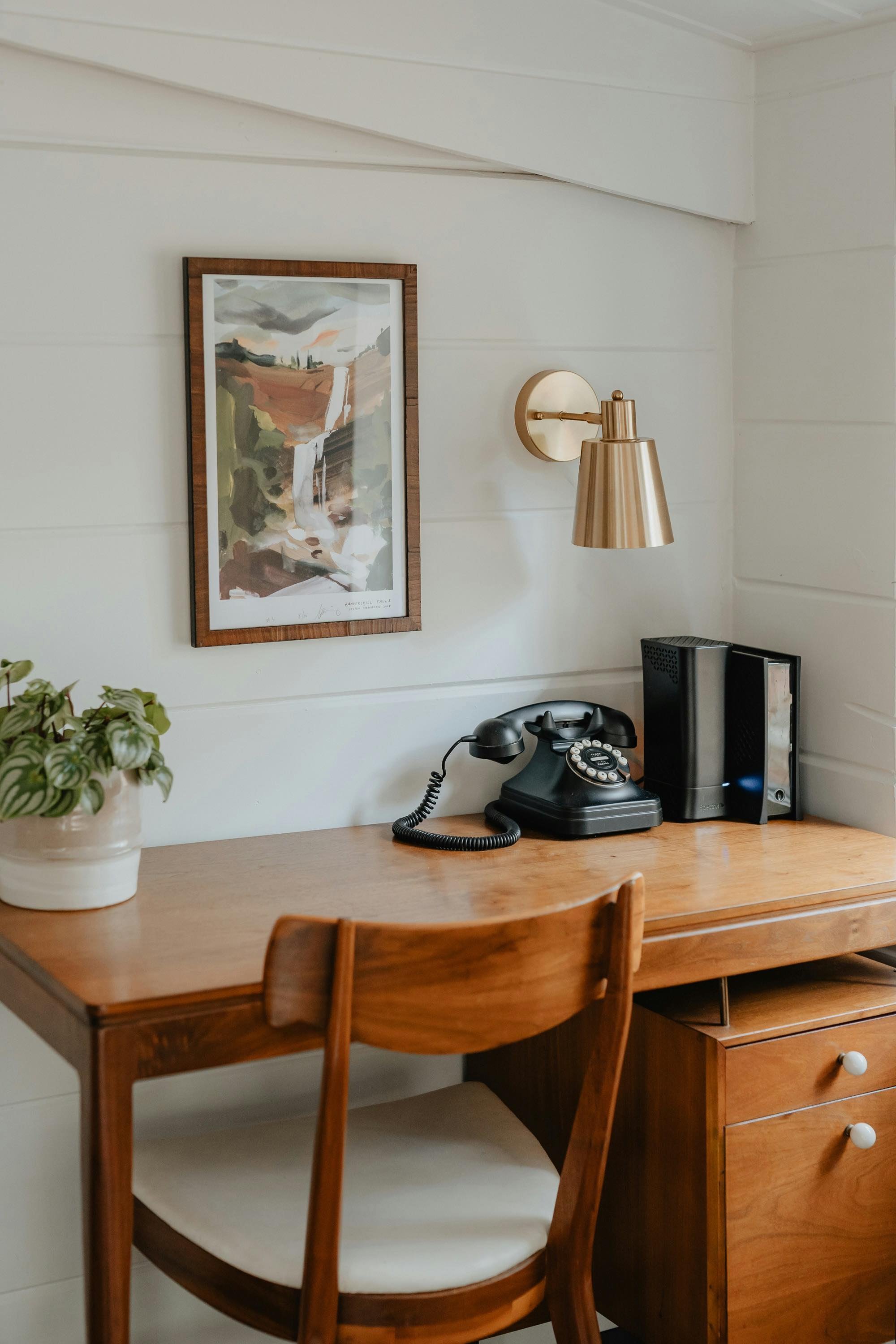 A wooden desk with a white cushioned chair, a potted plant, a vintage rotary phone, and a desktop computer with a monitor and speaker, against a white wall with a framed landscape painting and a gold wall-mounted lamp.