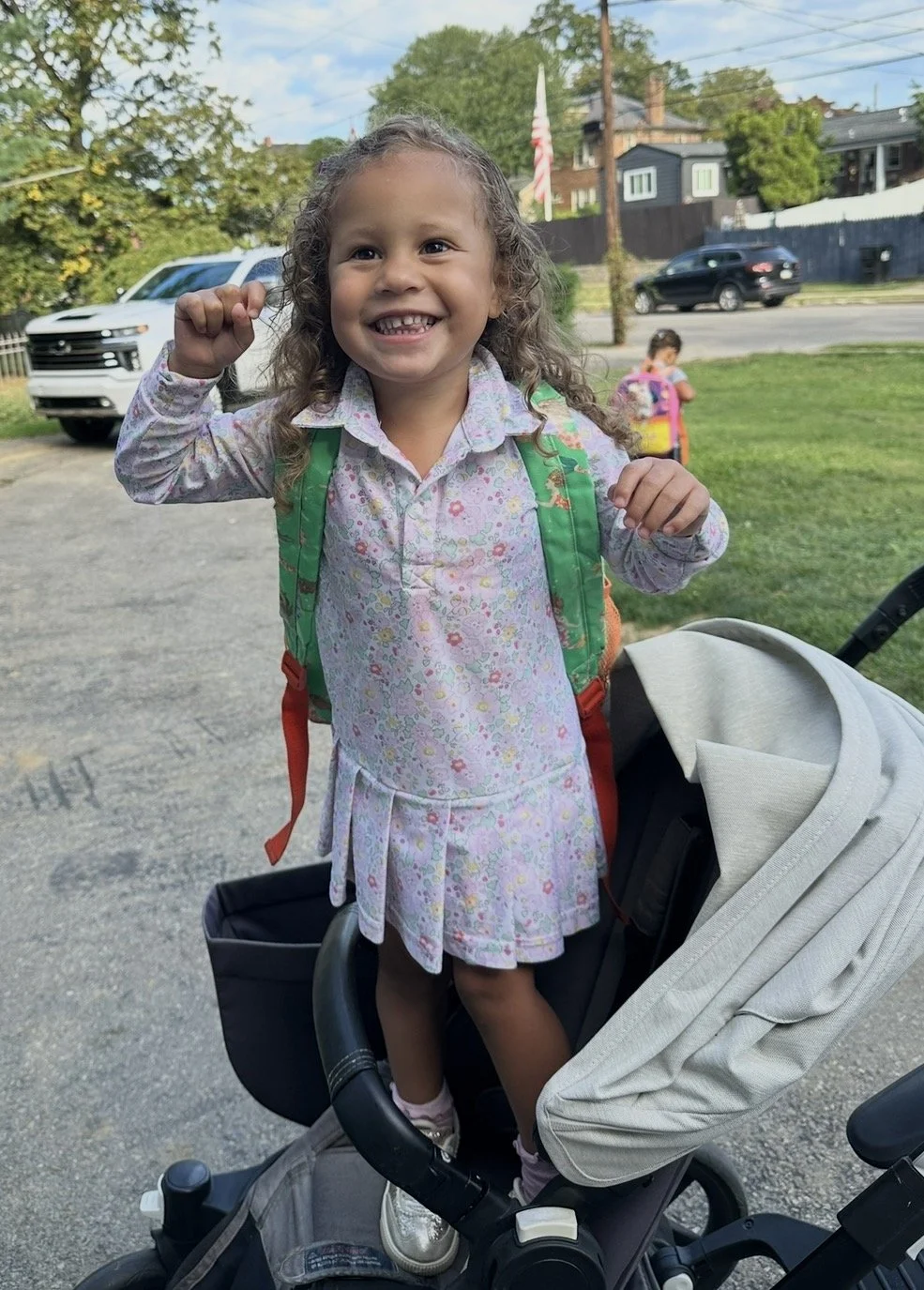 A young girl with curly hair smiling and showing a fist pump, standing in a stroller with a backpack, outdoors on a grassy area with trees and houses in the background.