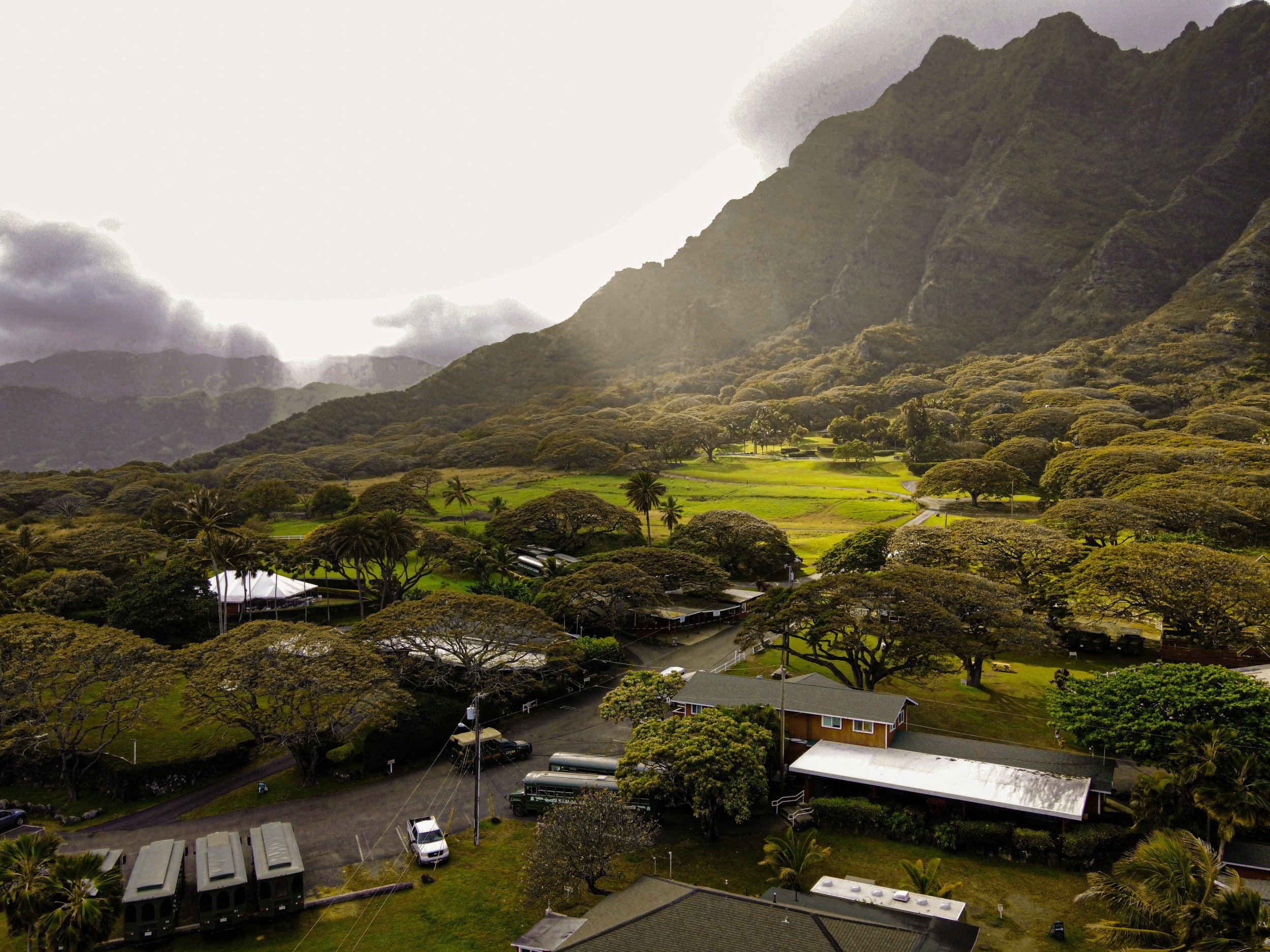 A scenic landscape with lush green trees, a mountain range in the background, and a clear sky with some clouds. There are buildings and vehicles in the foreground.