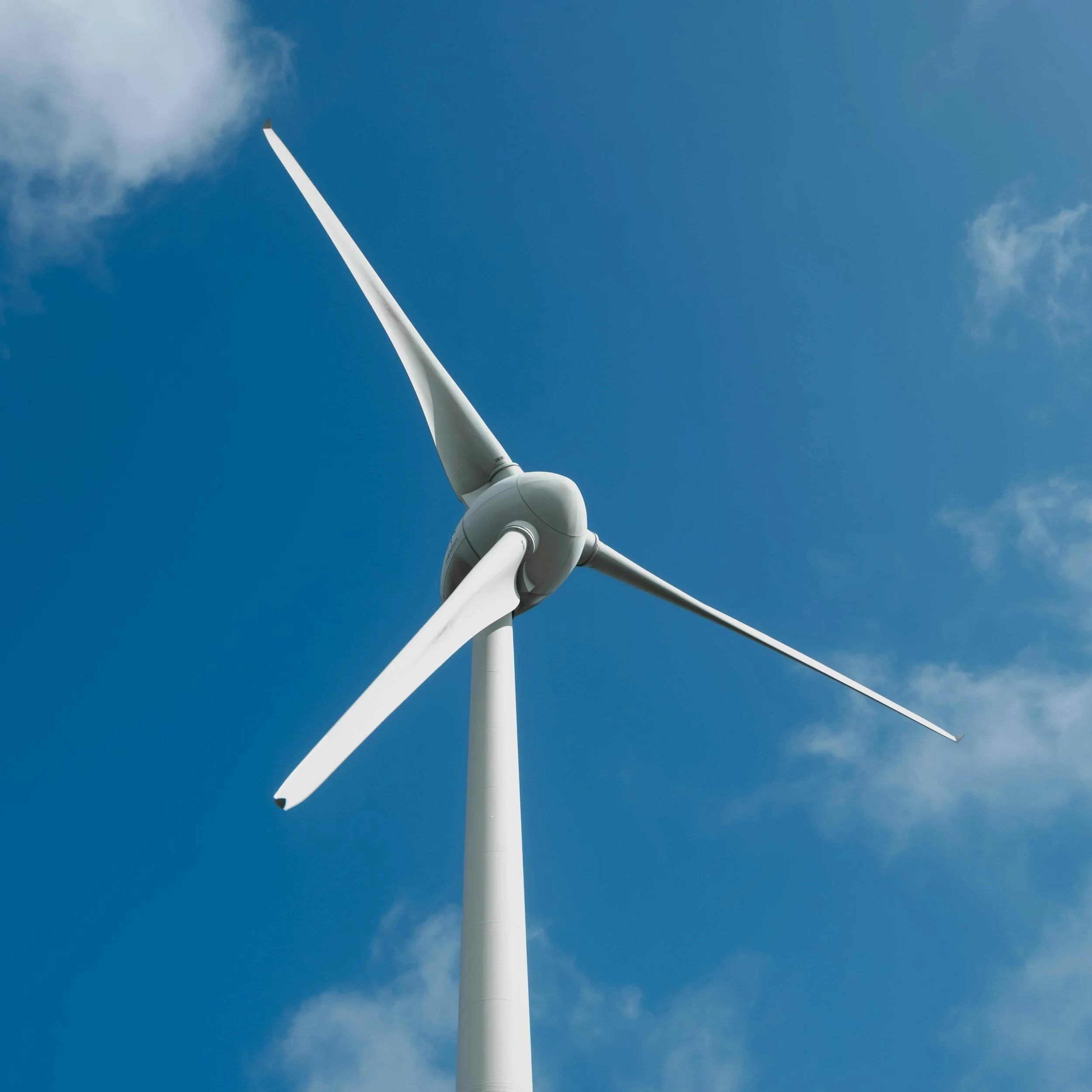 A close-up view looking up at a wind turbine set against a blue sky with some white clouds.