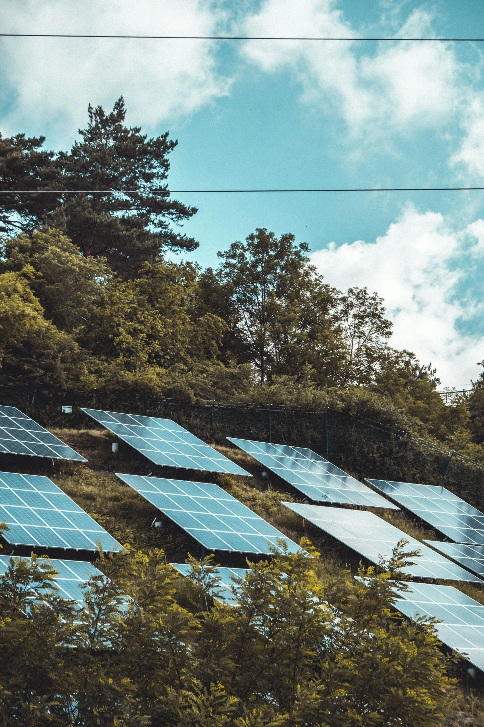 Solar panels installed on a hillside with trees and a partly cloudy sky in the background.