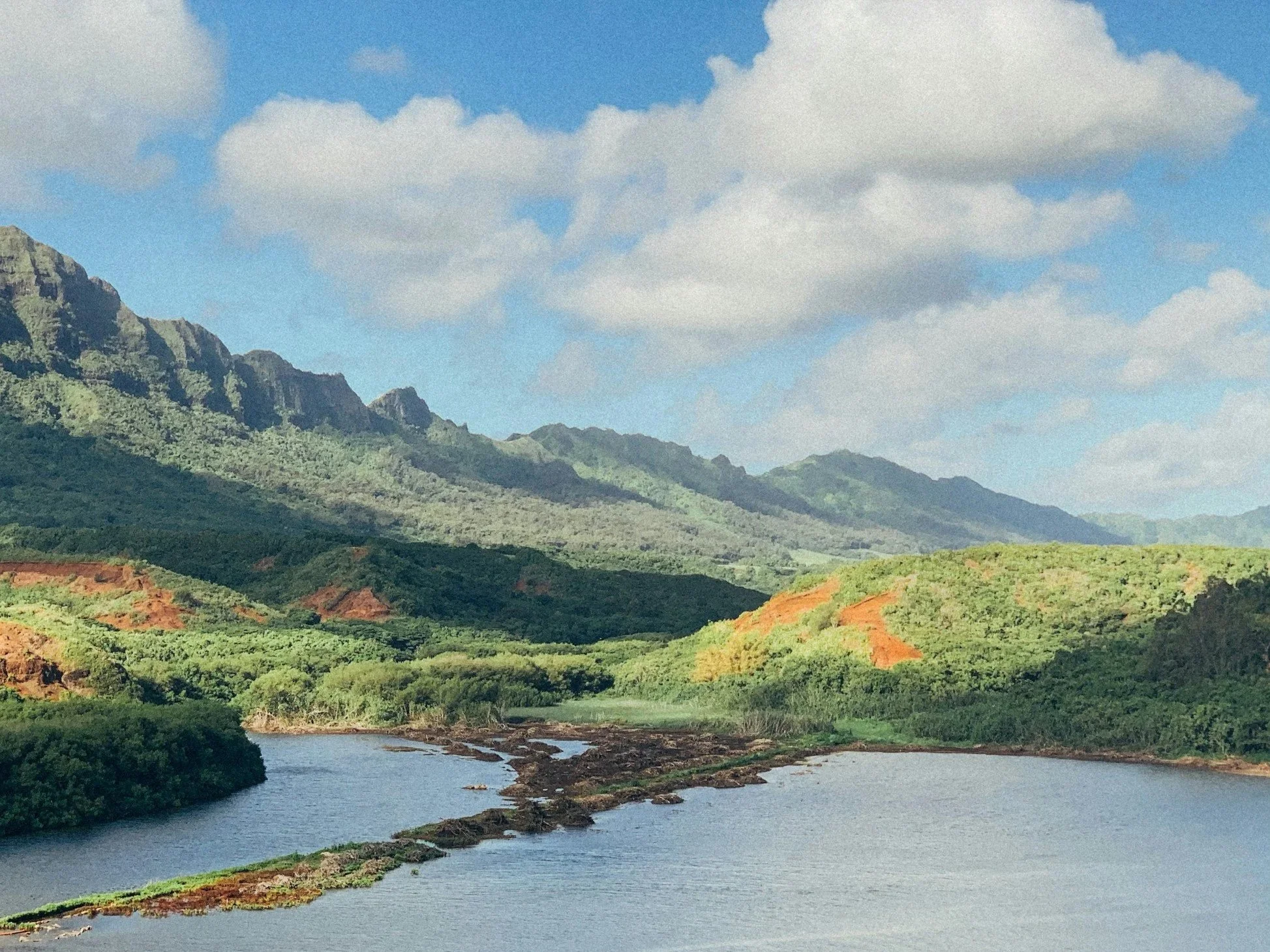 A scenic landscape of lush green mountains, a river flowing through the valley, clouds in the sky, and a mix of bright green and reddish soil on the terrain.