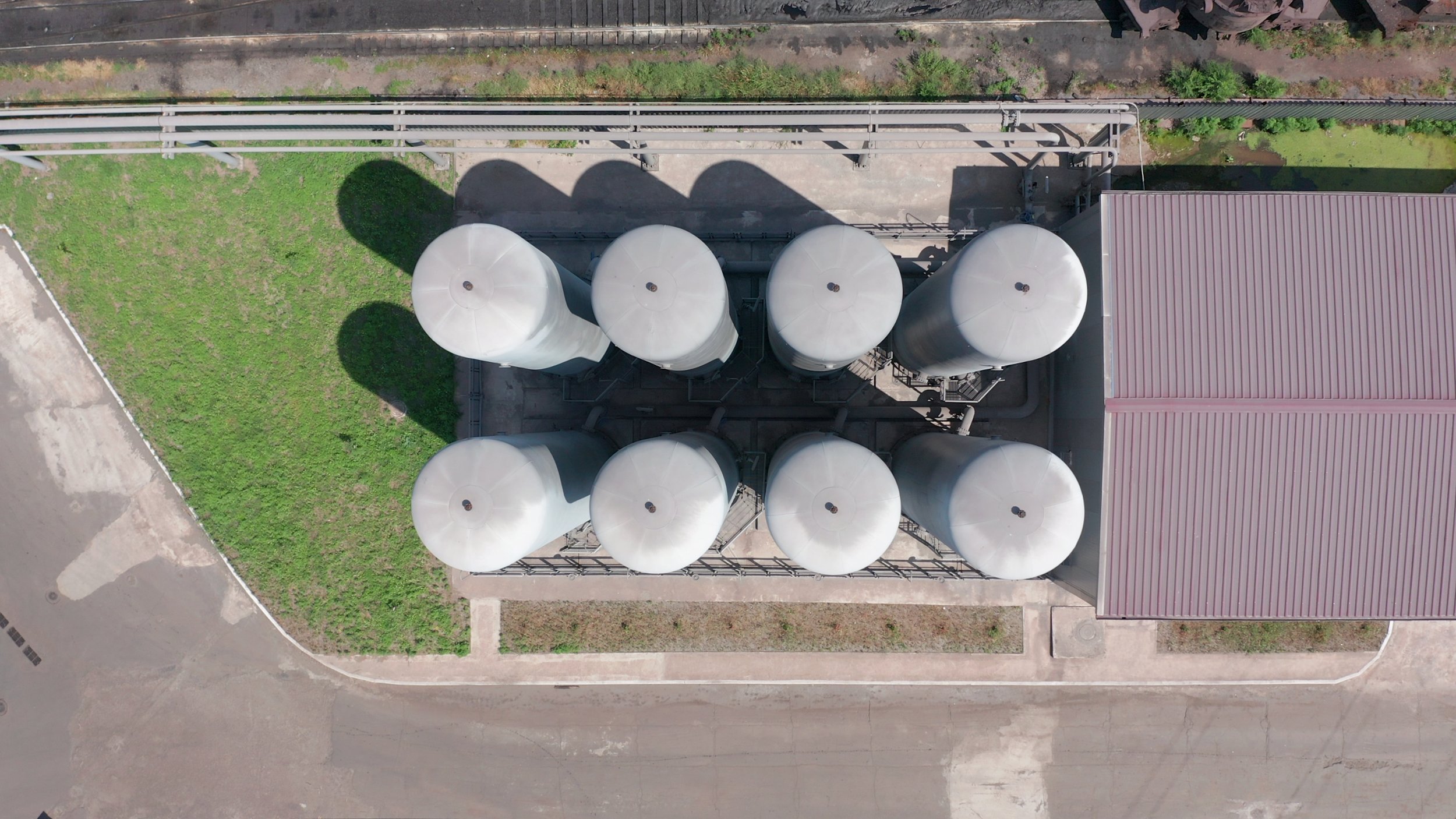 An aerial view of a building with seven large white cylindrical tanks on the roof, a red metal roof section, a patch of green grass, and paved surfaces surrounding the building.