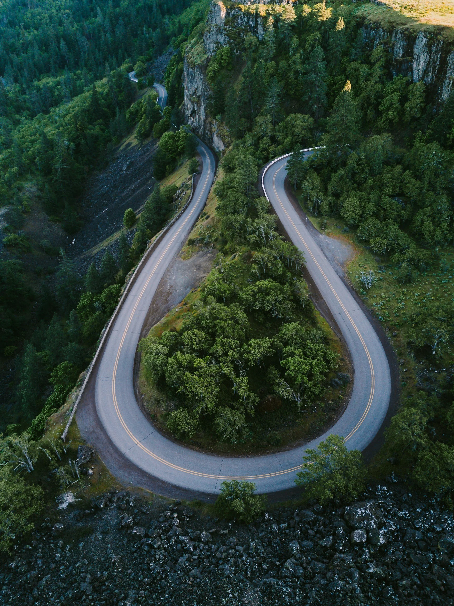 A winding mountain road in Rowena, Oregon surrounded by dense green forest, with steep cliffs and rocky terrain.