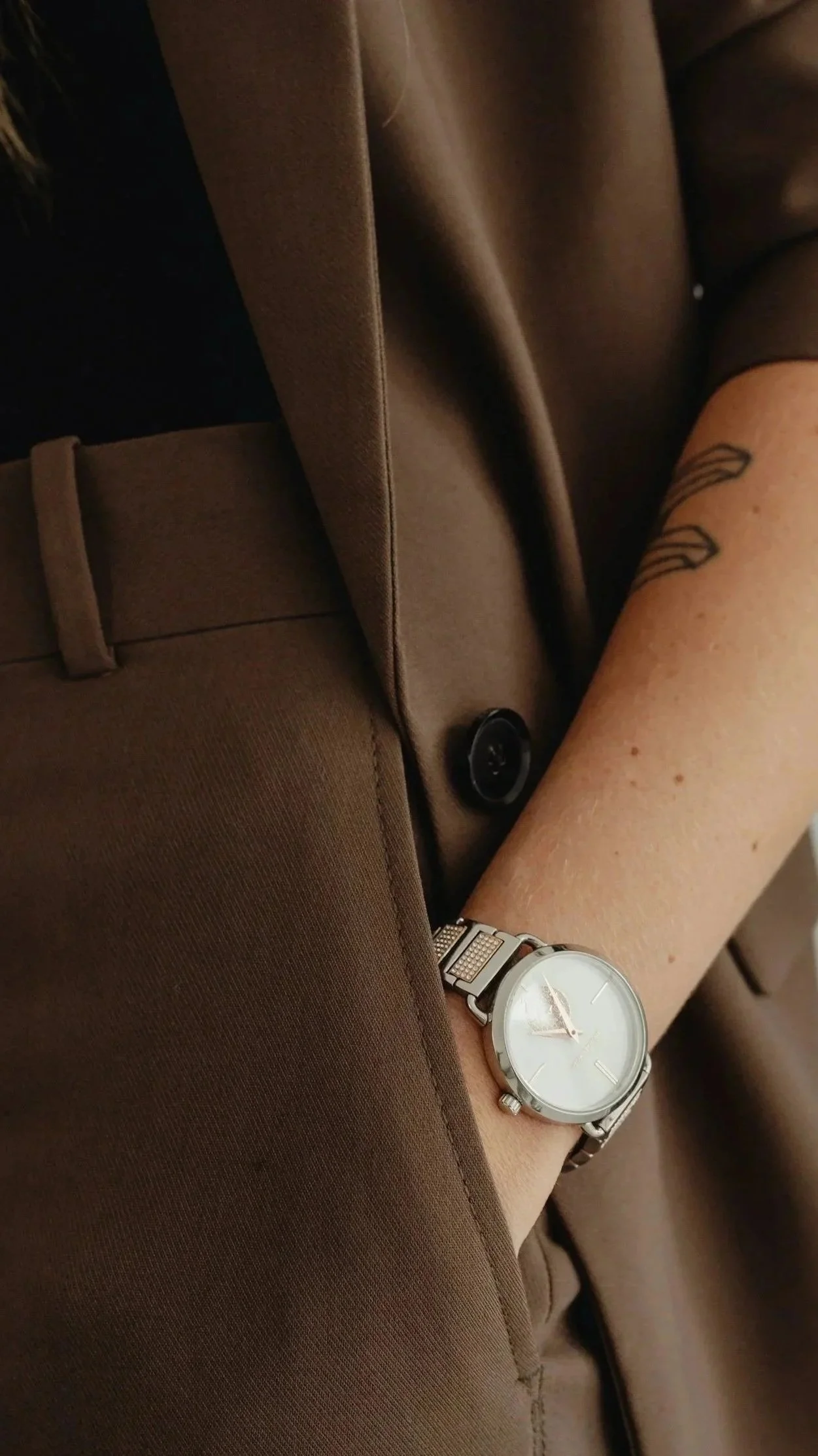 Close-up of a person's arm with a tattoo, wearing a wristwatch, with her hand in her pocket