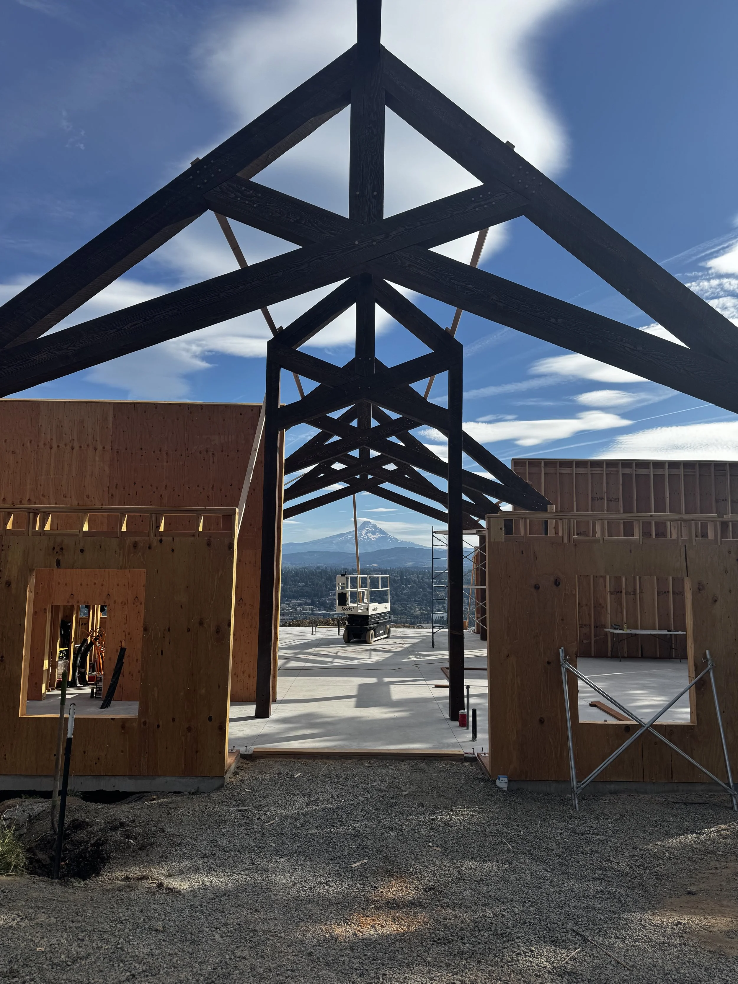 Construction site with wooden framing, wooden trusses and steel beams, Mount Hood in the background under partly cloudy sky.