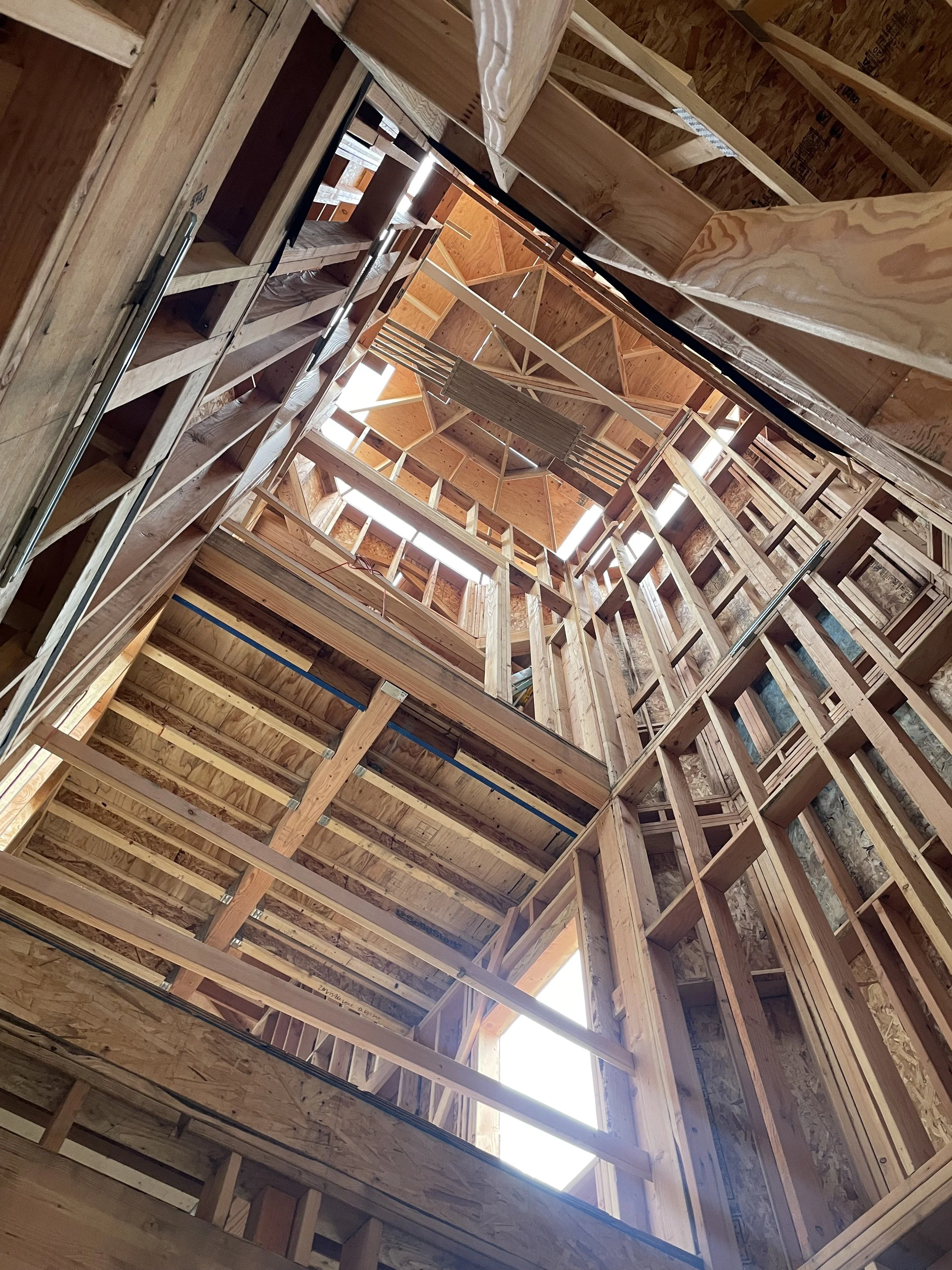 Inside view of a multi-story building under construction, showing exposed wooden framing and roof structure with sunlight coming through openings.