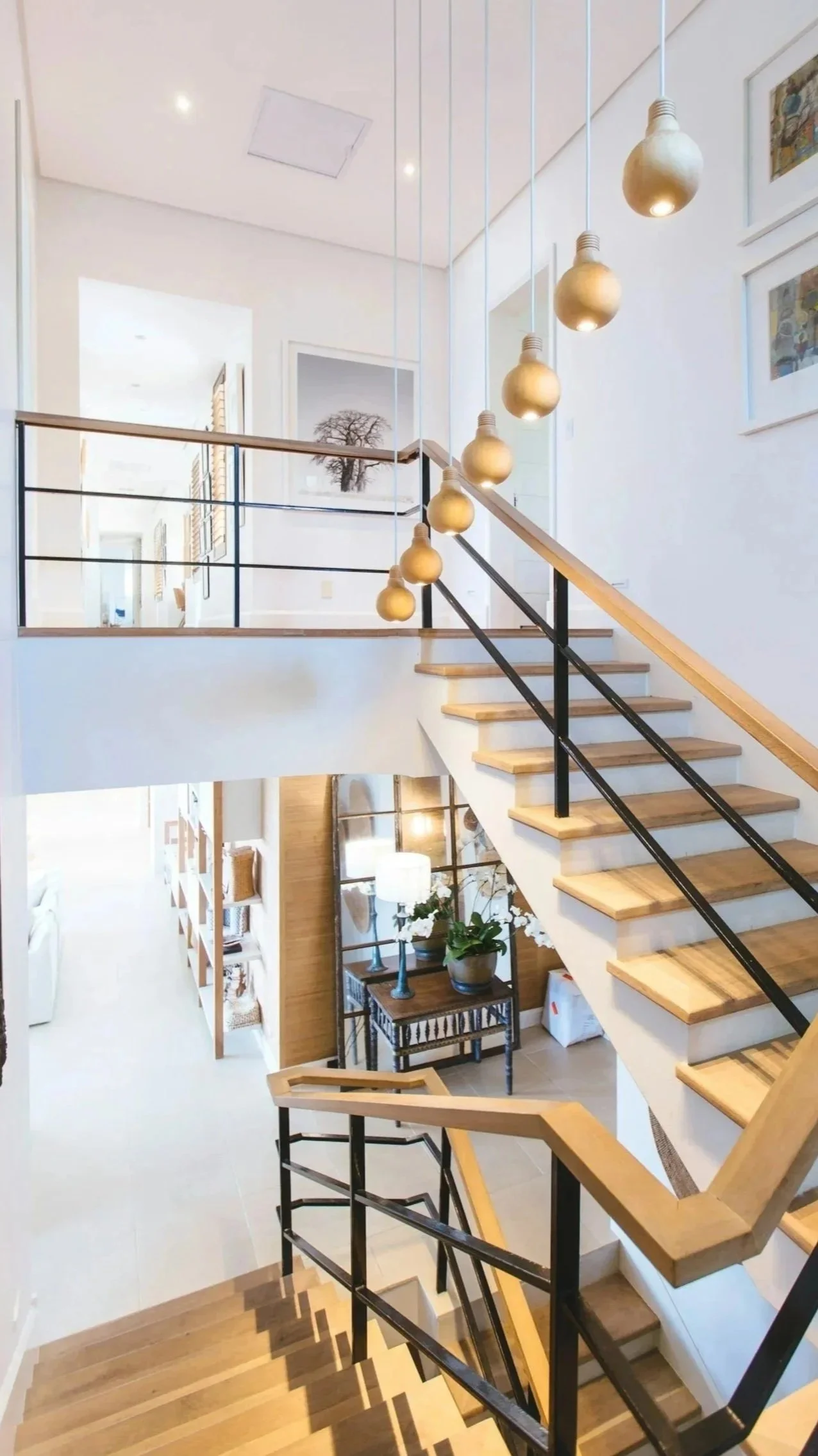Interior view of a house staircase with hanging light bulbs, black and wood handrails, and a living area with white walls and modern decor.