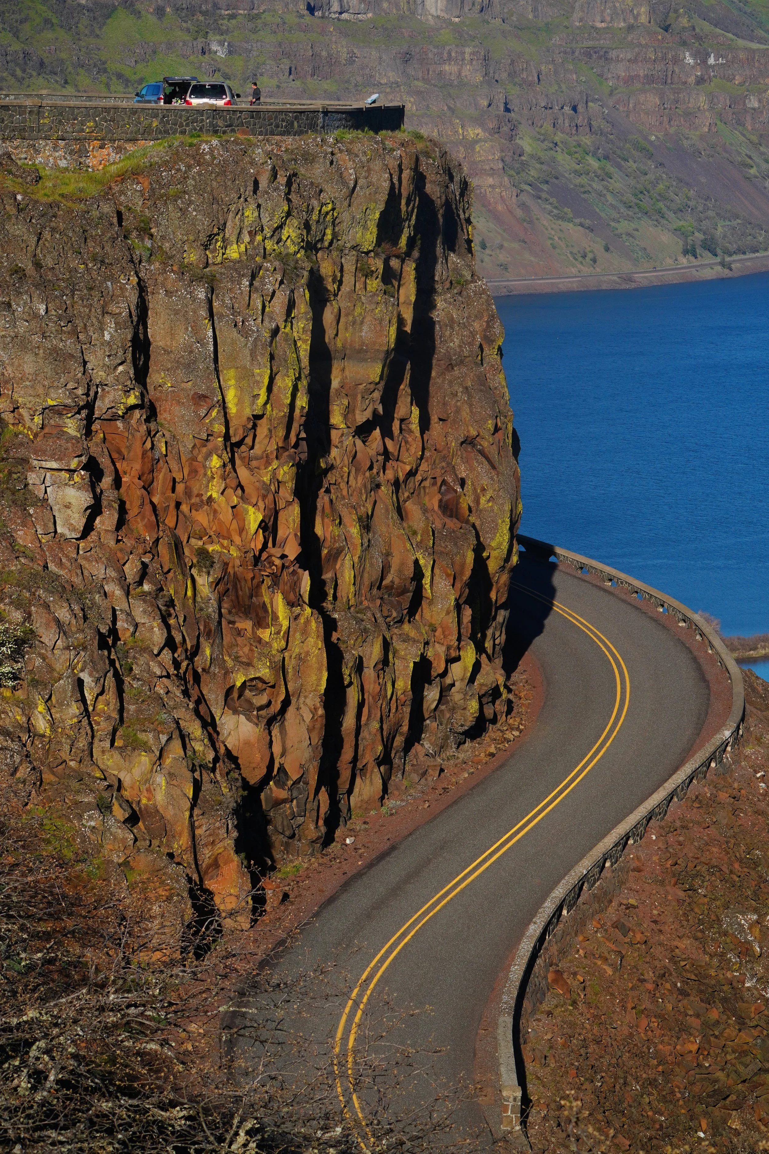 A winding road with a double yellow line travels along the edge of a rocky cliff overlooking the ocean. There are two cars parked on the top of the cliff and a person standing nearby. The landscape features rugged cliffs, the ocean below, and a hillside in the background.