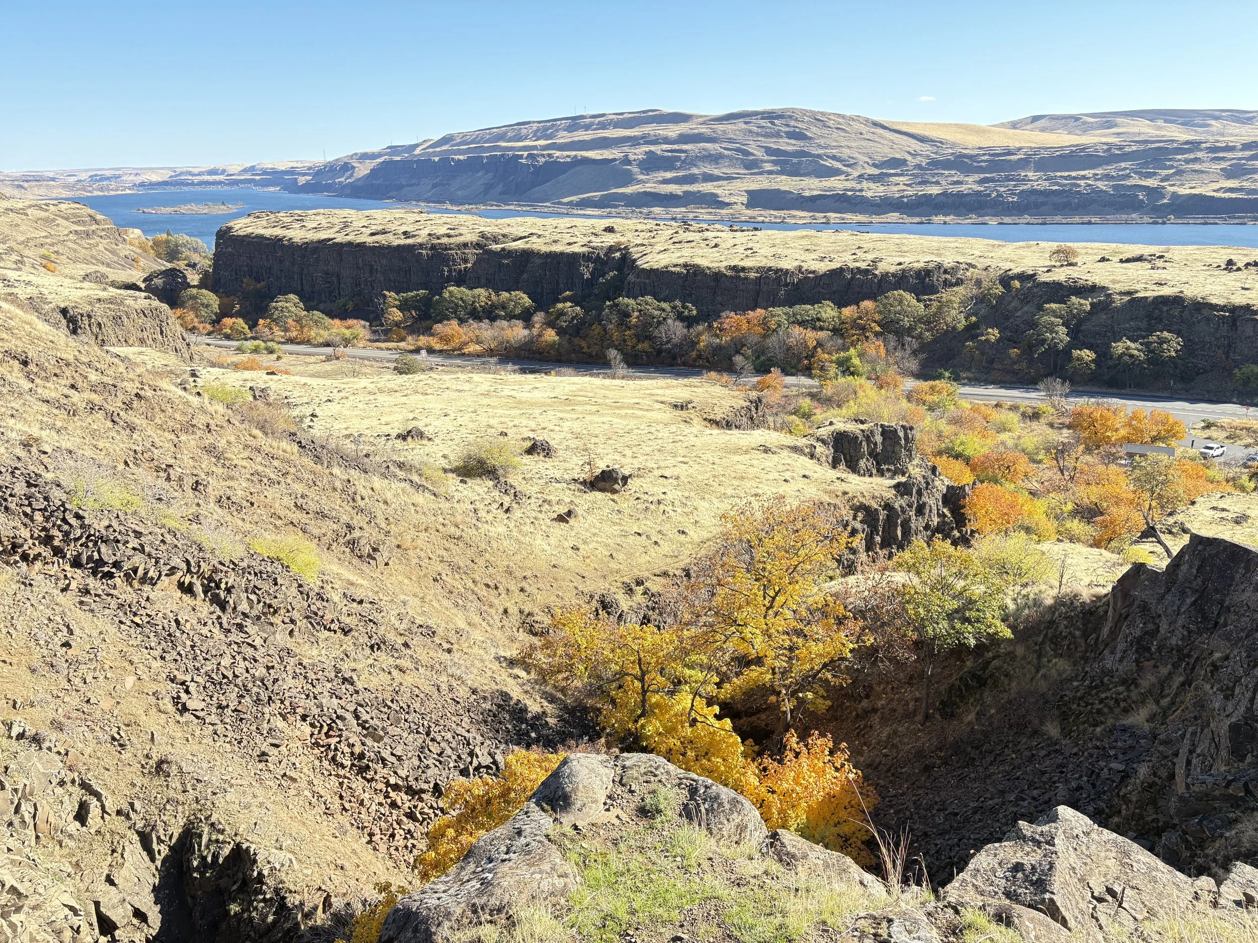 A scenic landscape of a river running through rocky hills with autumn-colored trees along the banks, and mountainous terrain in the background under a clear blue sky.